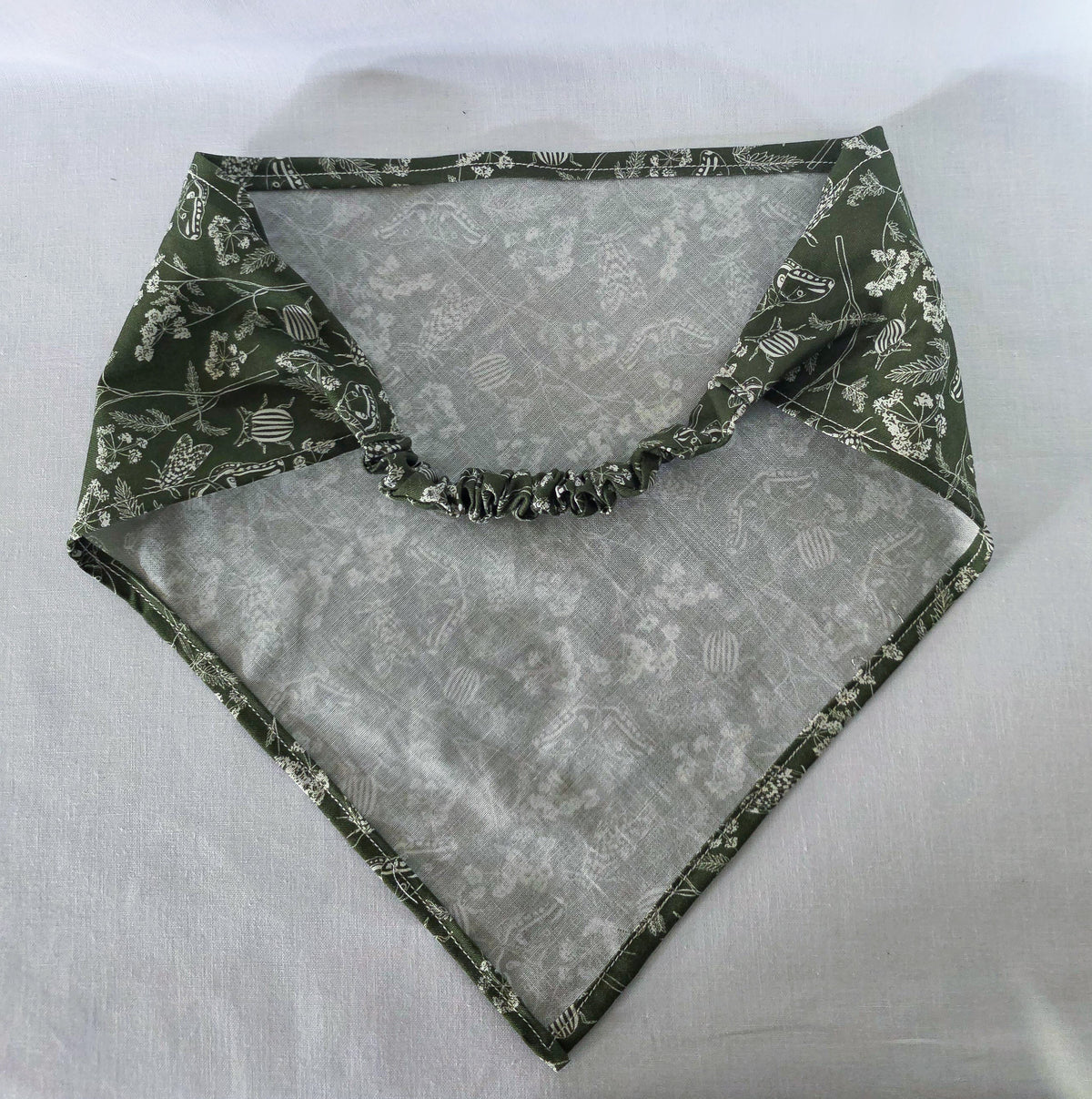 A green elastic hair bandana with a white floral and bug print on it. Laid on a white fabric background. 