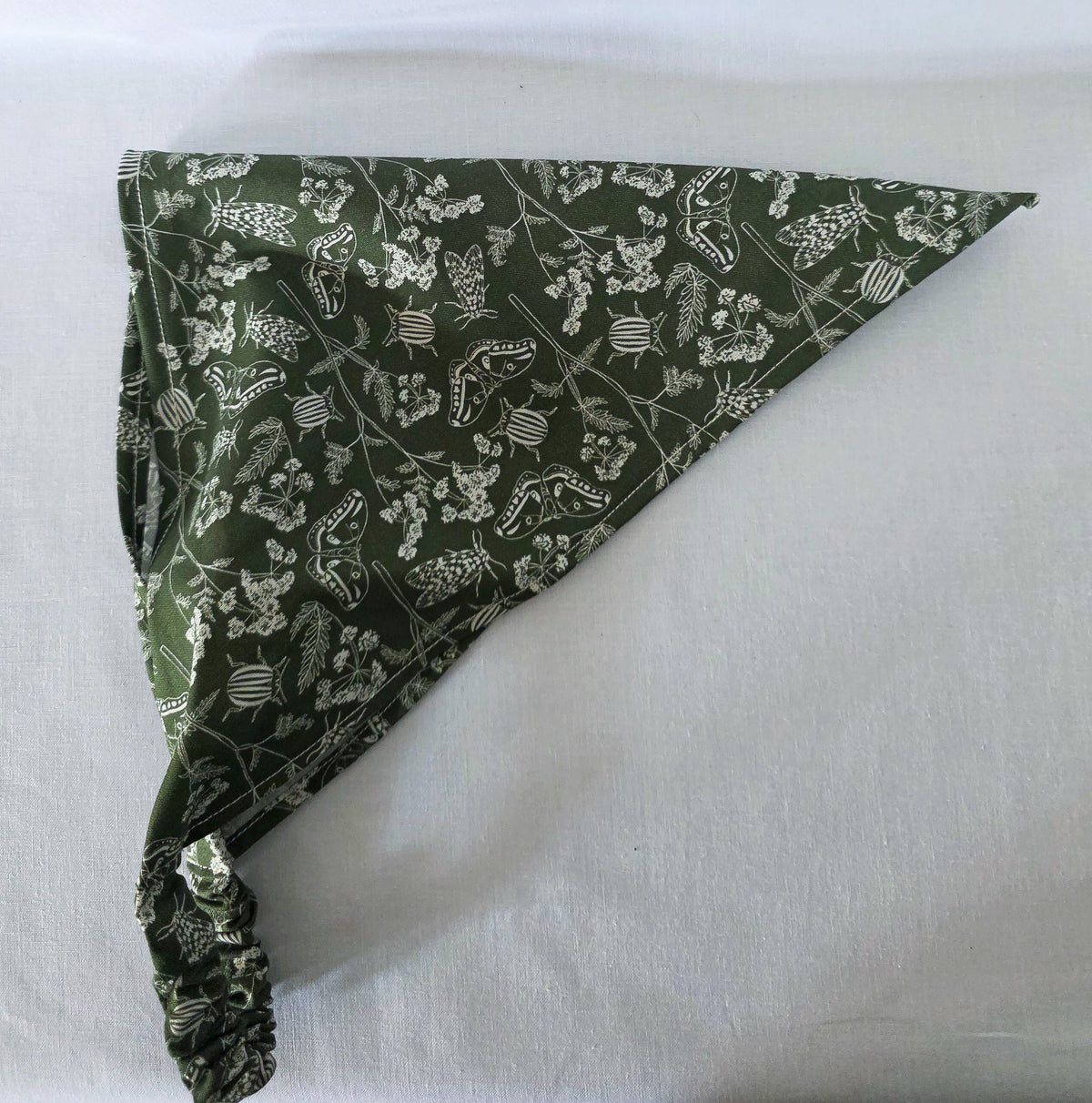 A green elastic hair bandana with a white floral and bug print on it. Laid flat on its side on a white fabric background. 