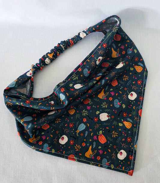 A fabric hair bandana laid flat on a light fabric background. It has an elastic band and a dark teal base covered in a colorful autumn floral print with pumpkins, leaves, and flowers.