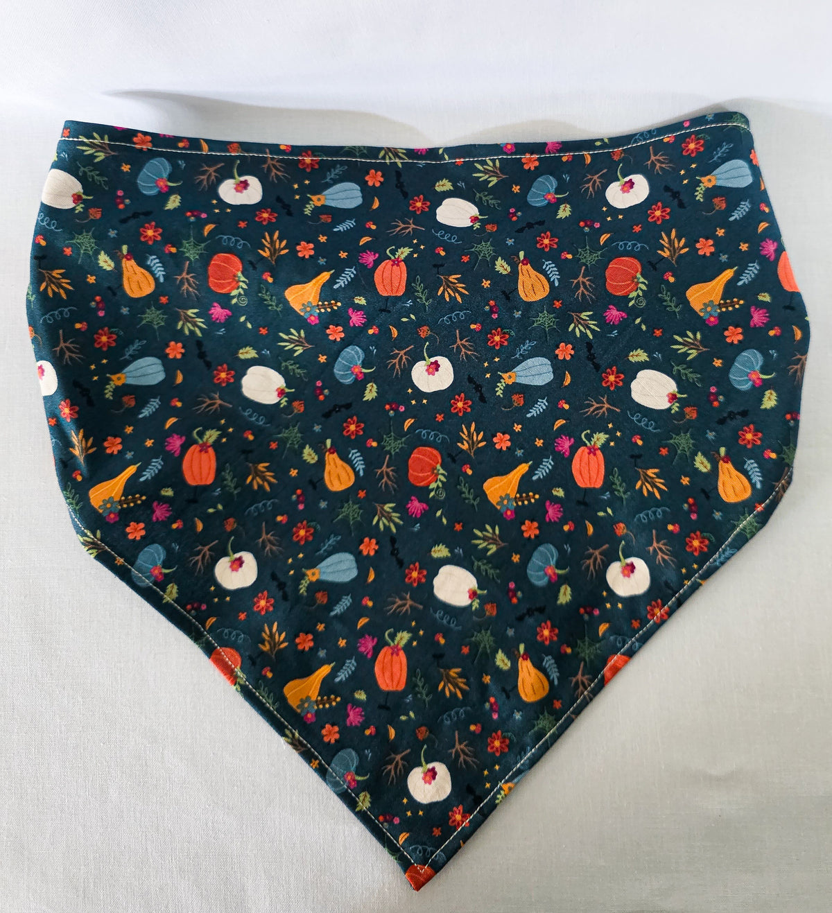 A fabric hair bandana laid flat on a light fabric background. It has an elastic band and a dark teal base covered in a colorful autumn floral print with pumpkins, leaves, and flowers.