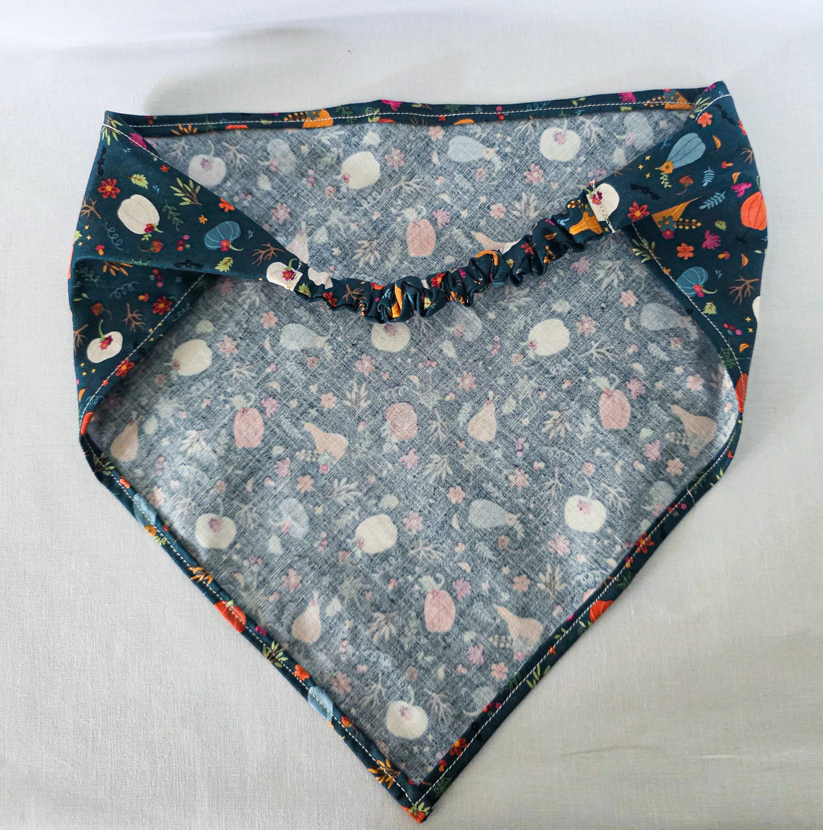 A fabric hair bandana laid flat on a light fabric background. It has an elastic band and a dark teal base covered in a colorful autumn floral print with pumpkins, leaves, and flowers.