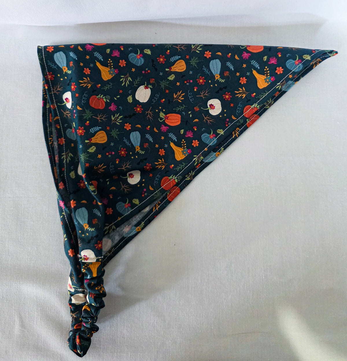 A fabric hair bandana laid flat on its side on a light fabric background. It has an elastic band and a dark teal base covered in a colorful autumn floral print with pumpkins, leaves, and flowers.