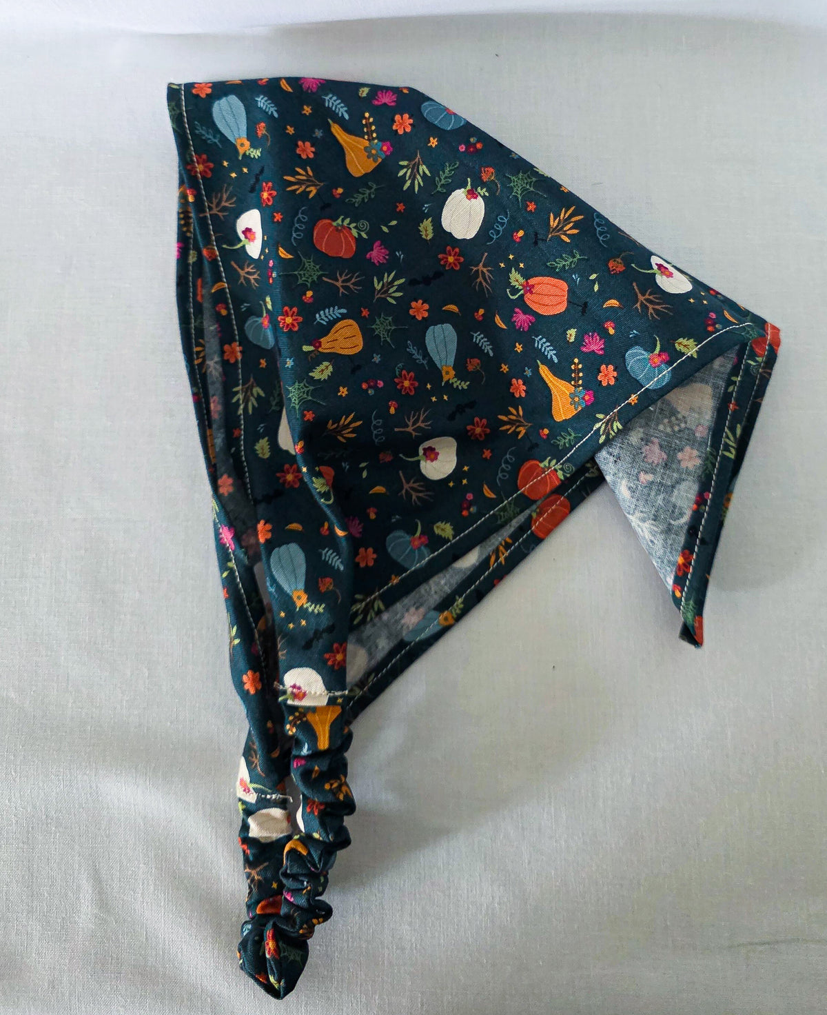 A fabric hair bandana laid flat draped on its side on a light fabric background. It has an elastic band and a dark teal base covered in a colorful autumn floral print with pumpkins, leaves, and flowers.