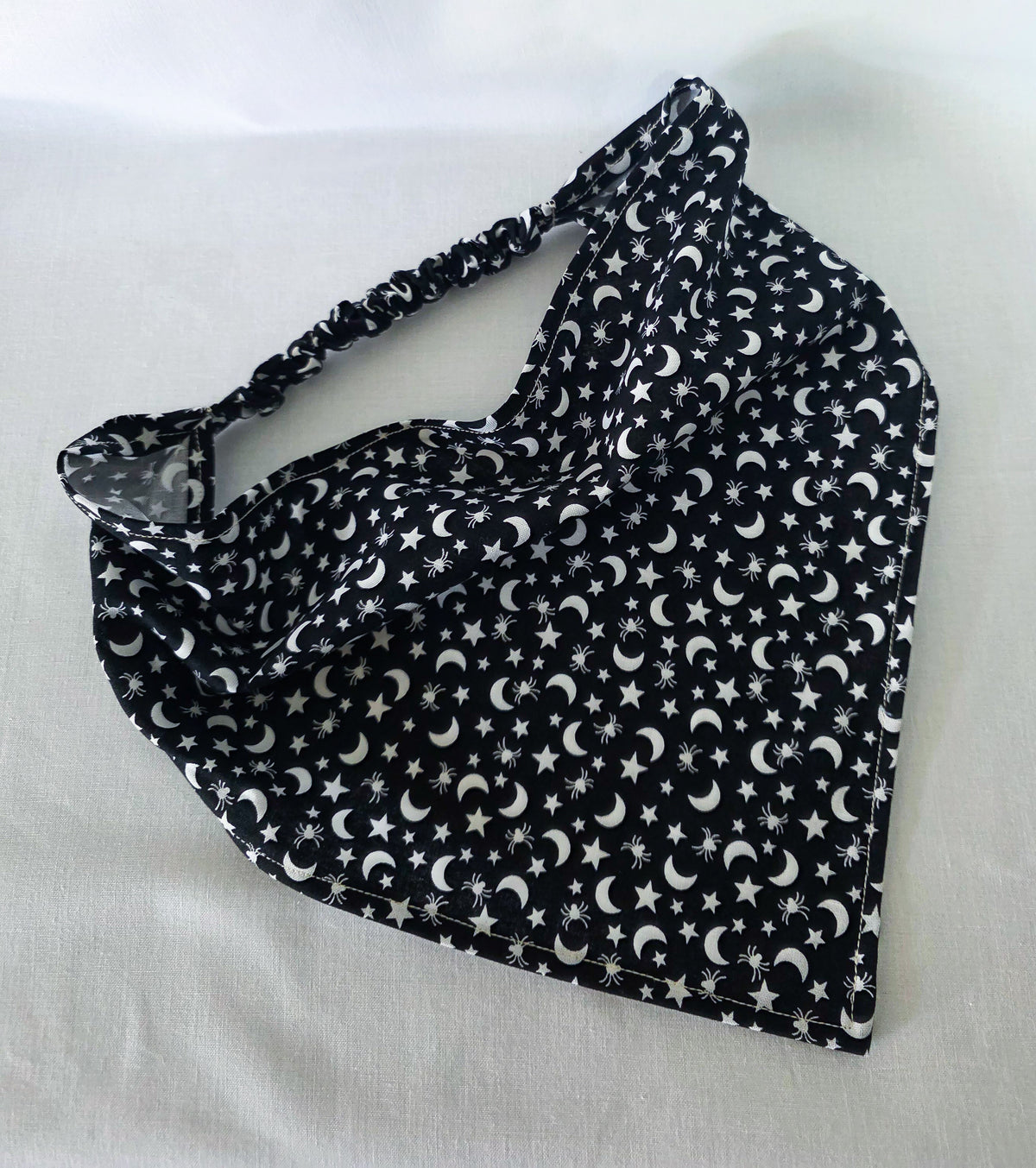 A black fabric elastic hair bandana with a white celestial pattern of crescent moons, stars, and spiders laid flat on a white fabric background.