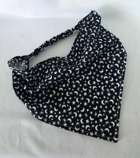 A black fabric elastic hair bandana with a white celestial pattern of crescent moons, stars, and spiders laid flat on a white fabric background.