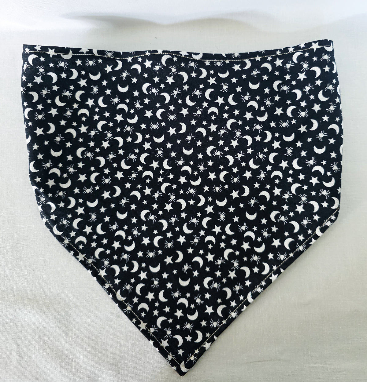A black fabric elastic hair bandana with a white celestial pattern of crescent moons, stars, and spiders laid flat on a white fabric background.