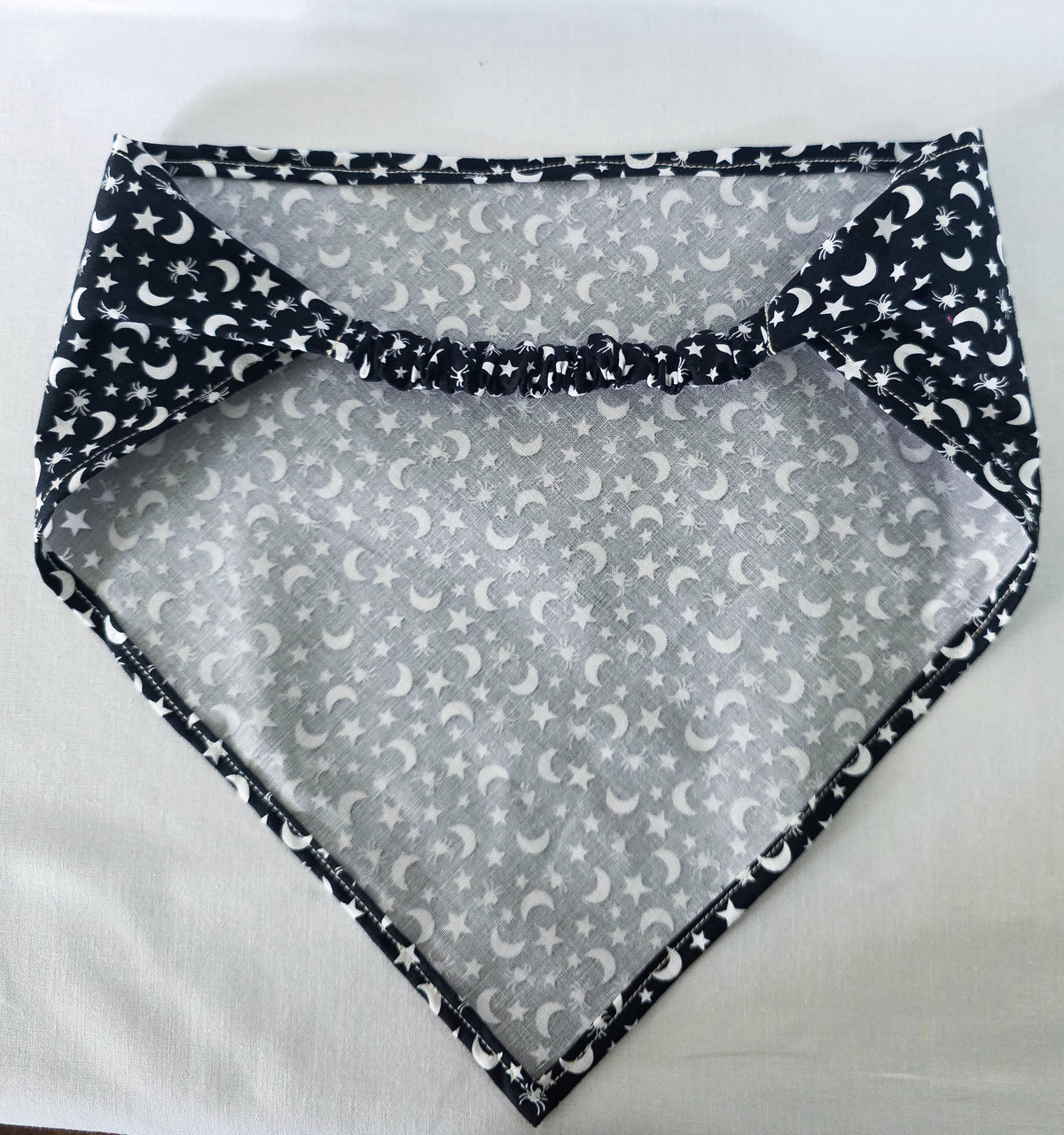 A black fabric elastic hair bandana with a white celestial pattern of crescent moons, stars, and spiders laid flat on a white fabric background.