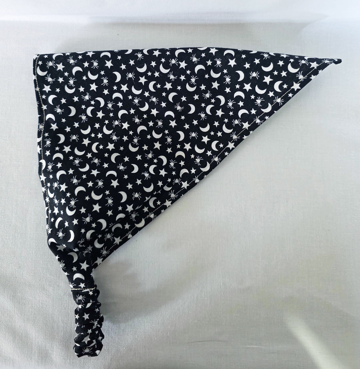 A black fabric elastic hair bandana with a white celestial pattern of crescent moons, stars, and spiders laid flat on its side on a white fabric background.