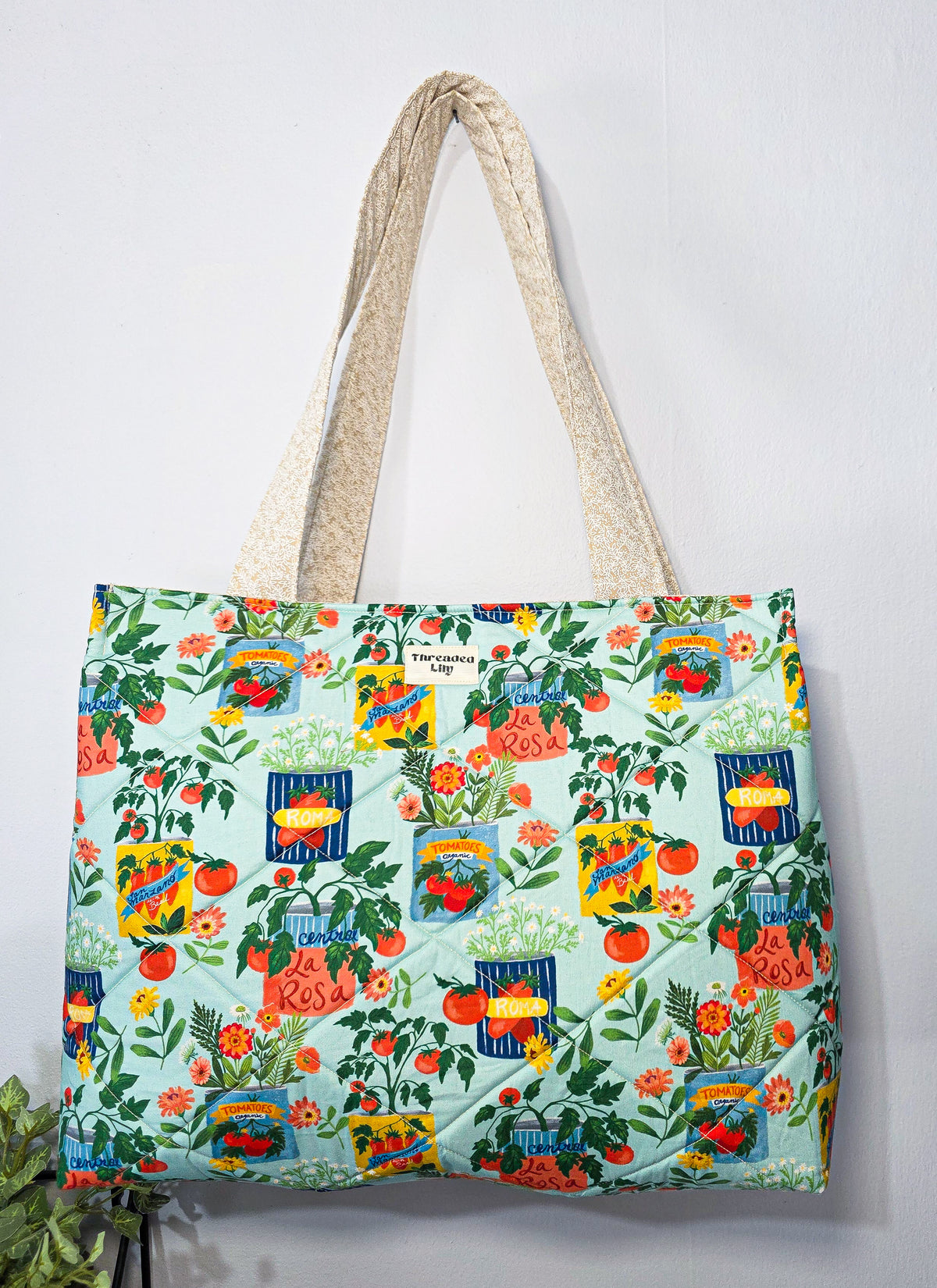 A quilted tote bag with a colorful garden and tomato print on a light mint green background, featuring cream-colored woven shoulder straps, hanging against a plain white wall.
