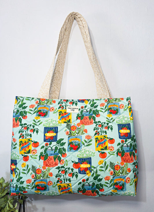 A quilted tote bag with a colorful garden and tomato print on a light mint green background, featuring cream-colored woven shoulder straps, hanging against a plain white wall.