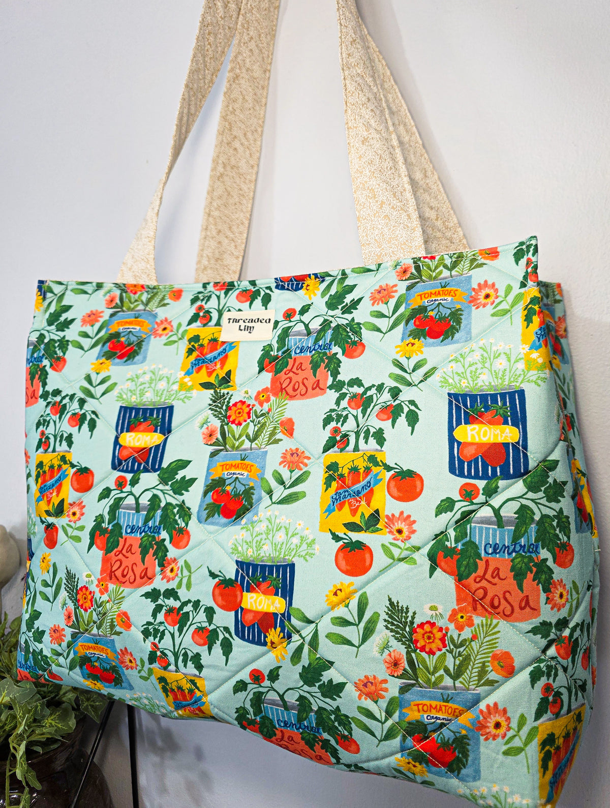 A quilted tote bag with a colorful garden print featuring tomatoes, herbs, flowers, and baskets, hanging against a white wall. The bag has a mint green base and cream-colored shoulder straps.