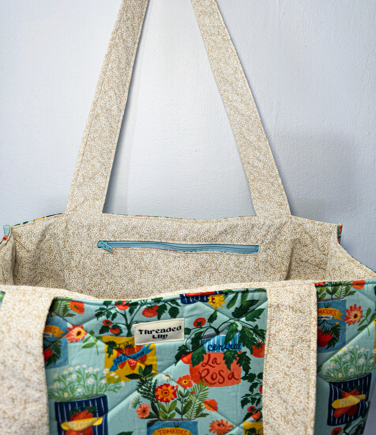 A close-up of a fabric tote bag with beige floral straps and trim, and a light blue floral front panel featuring botanical prints and tomato cans.