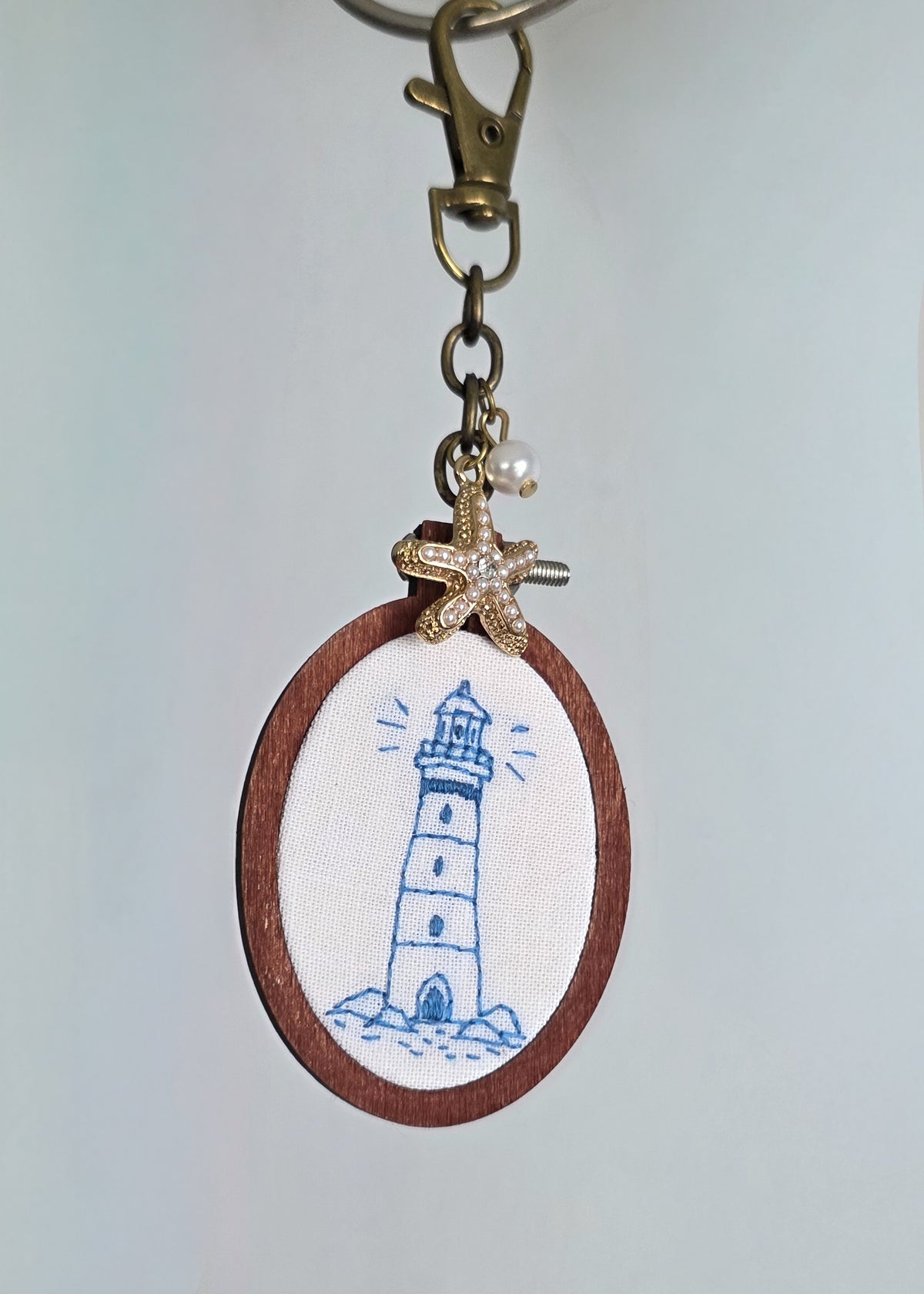 Hand embroidered keychain, featuring an outline of a lighthouse, done in blue thread. I has a starfish and pearl charm as well. It is hanging against a light background. 