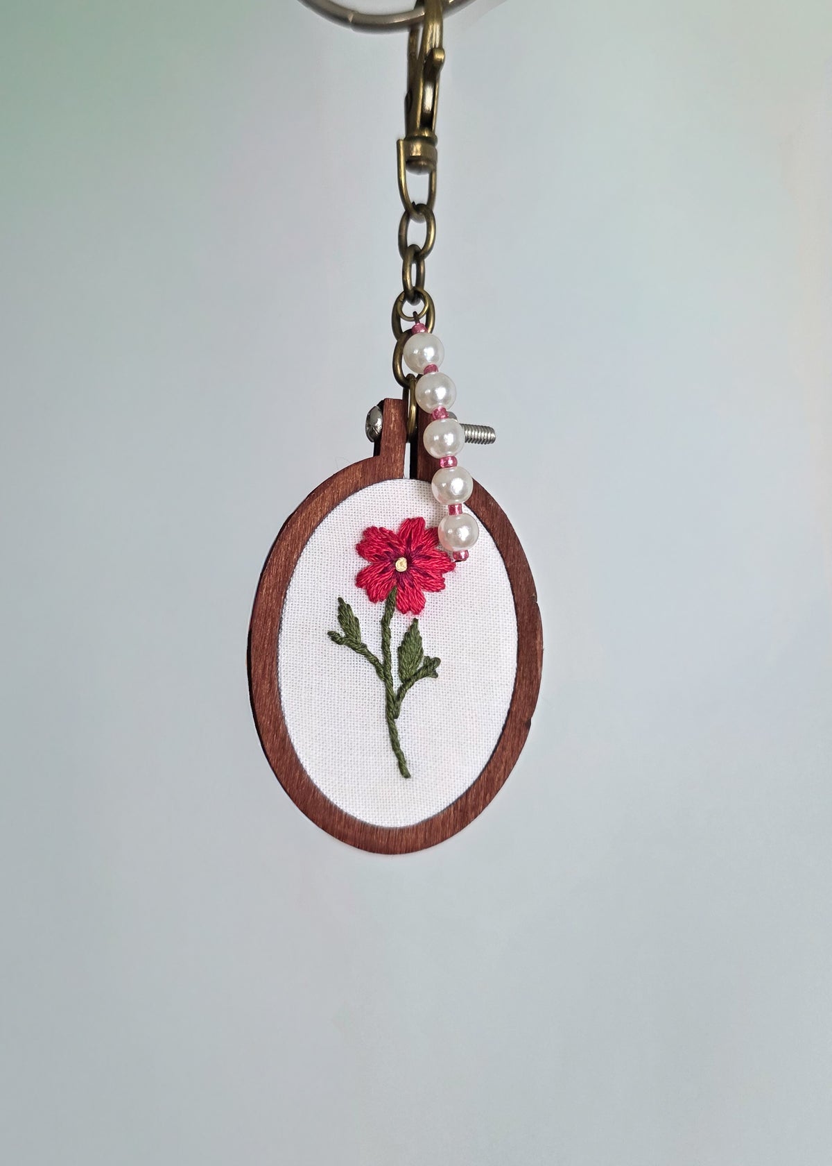 floral hand embroidered keychain, featuring a pink flower in a wooden hoop with pearl charms. Hanging against a light background. 