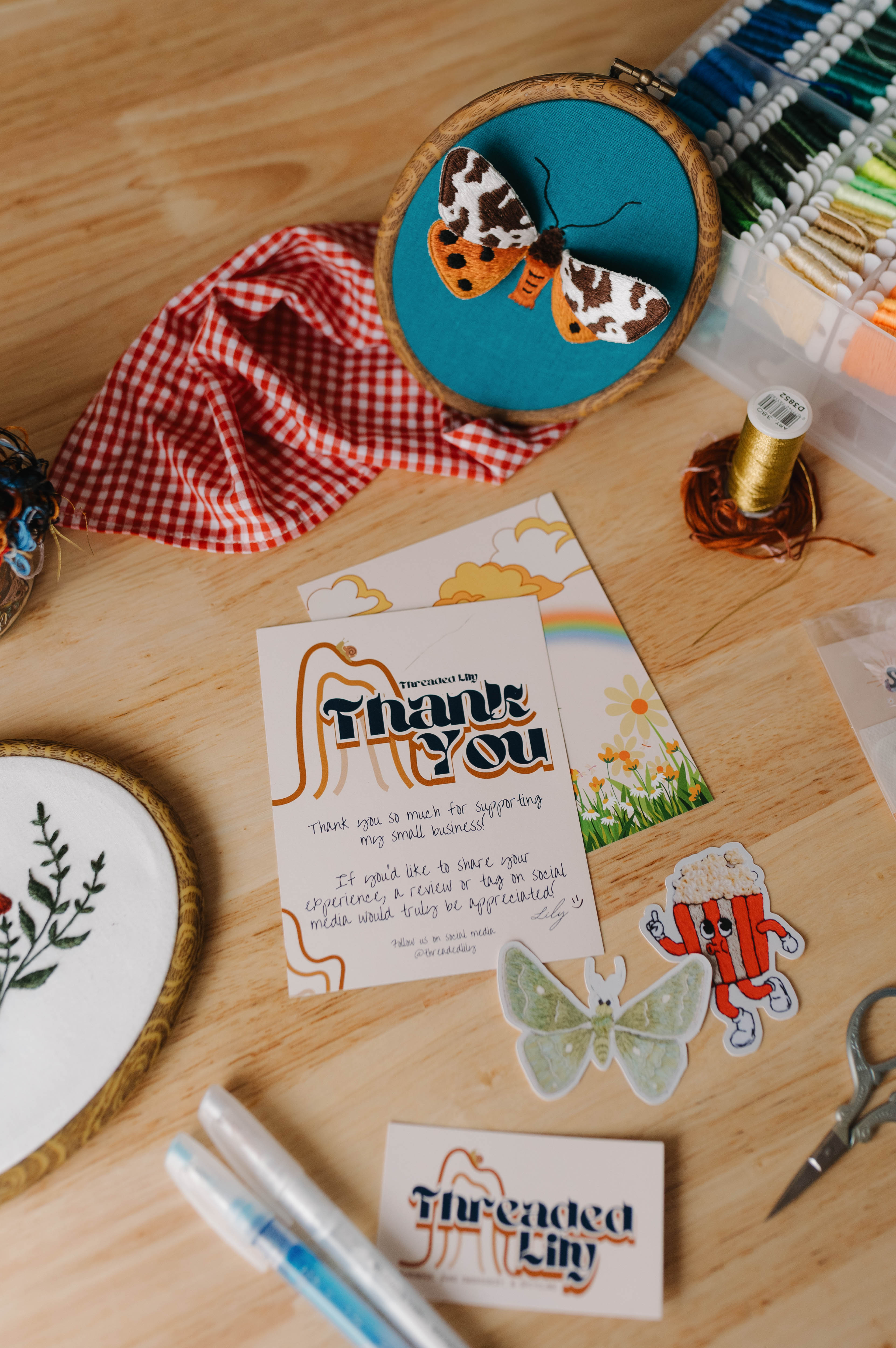 Embroidery hoop with embroidered items on a wooden table, surrounded by craft supplies.