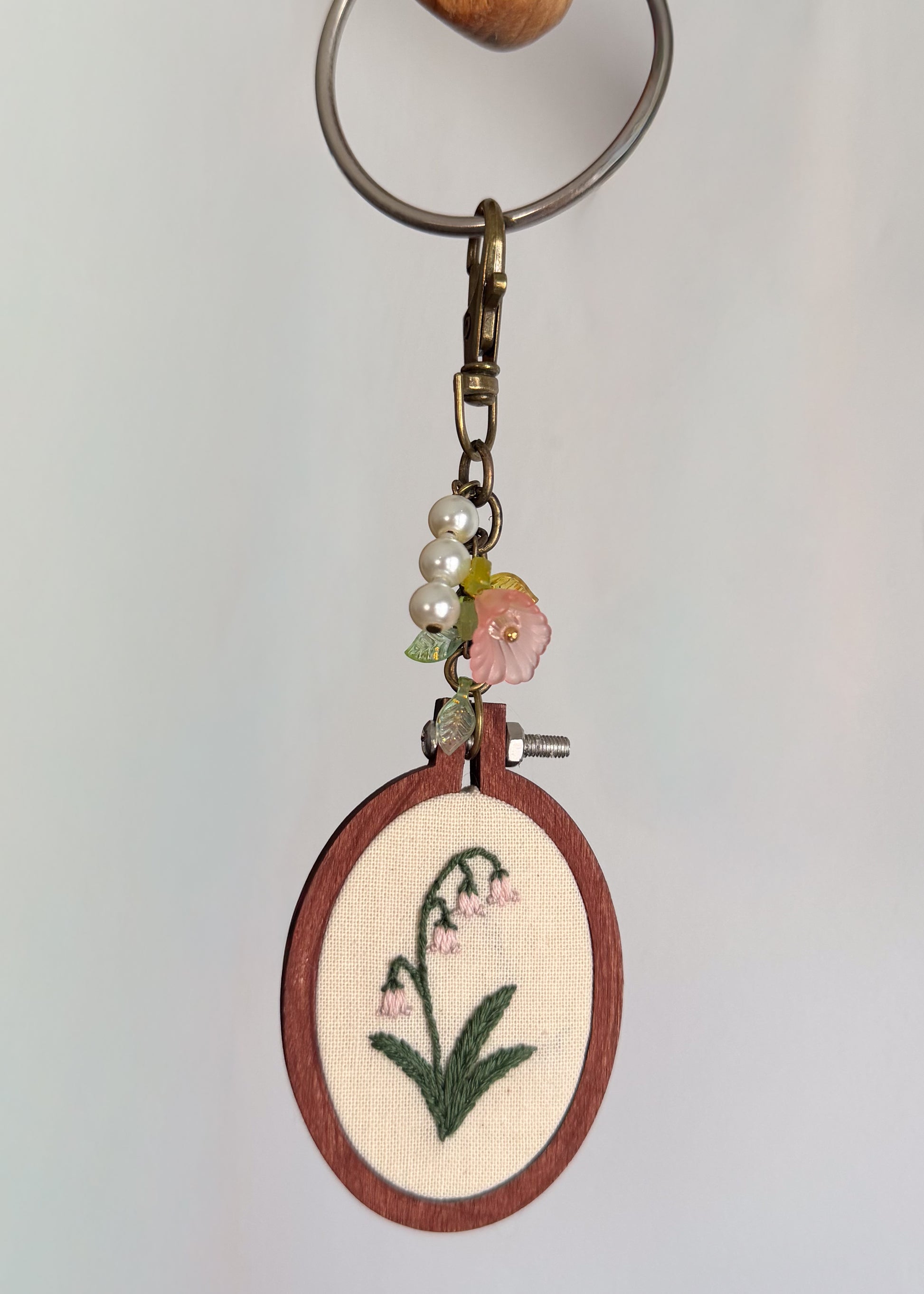A handmade keychain featuring an oval embroidery hoop with a lily of the valley design, hanging from a metal key ring with decorative pearl beads and a pink flower charm.