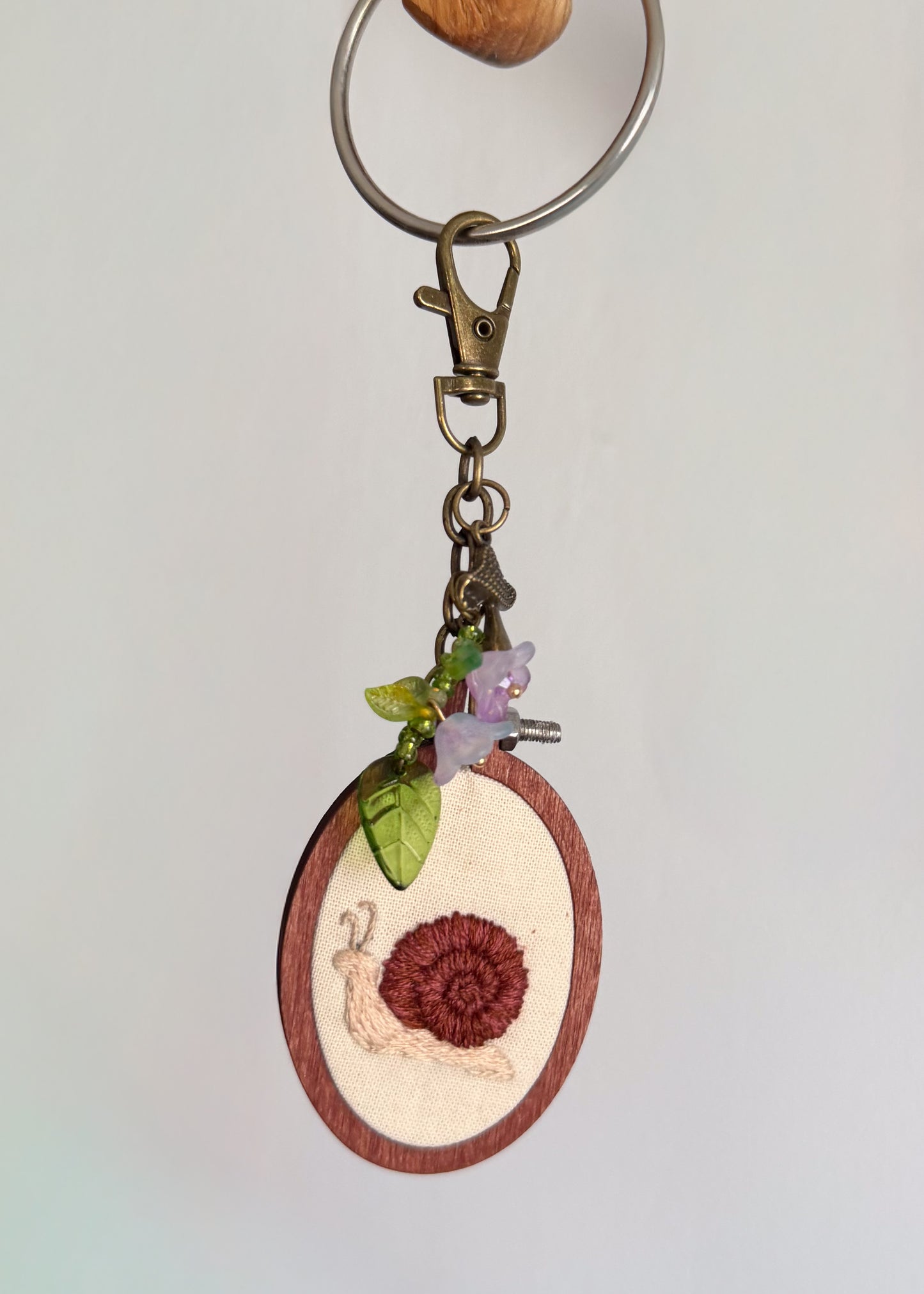 A close-up of a keychain featuring a small oval wooden charm with a snail design, hanging from a metal split ring and clasp against a plain light background.