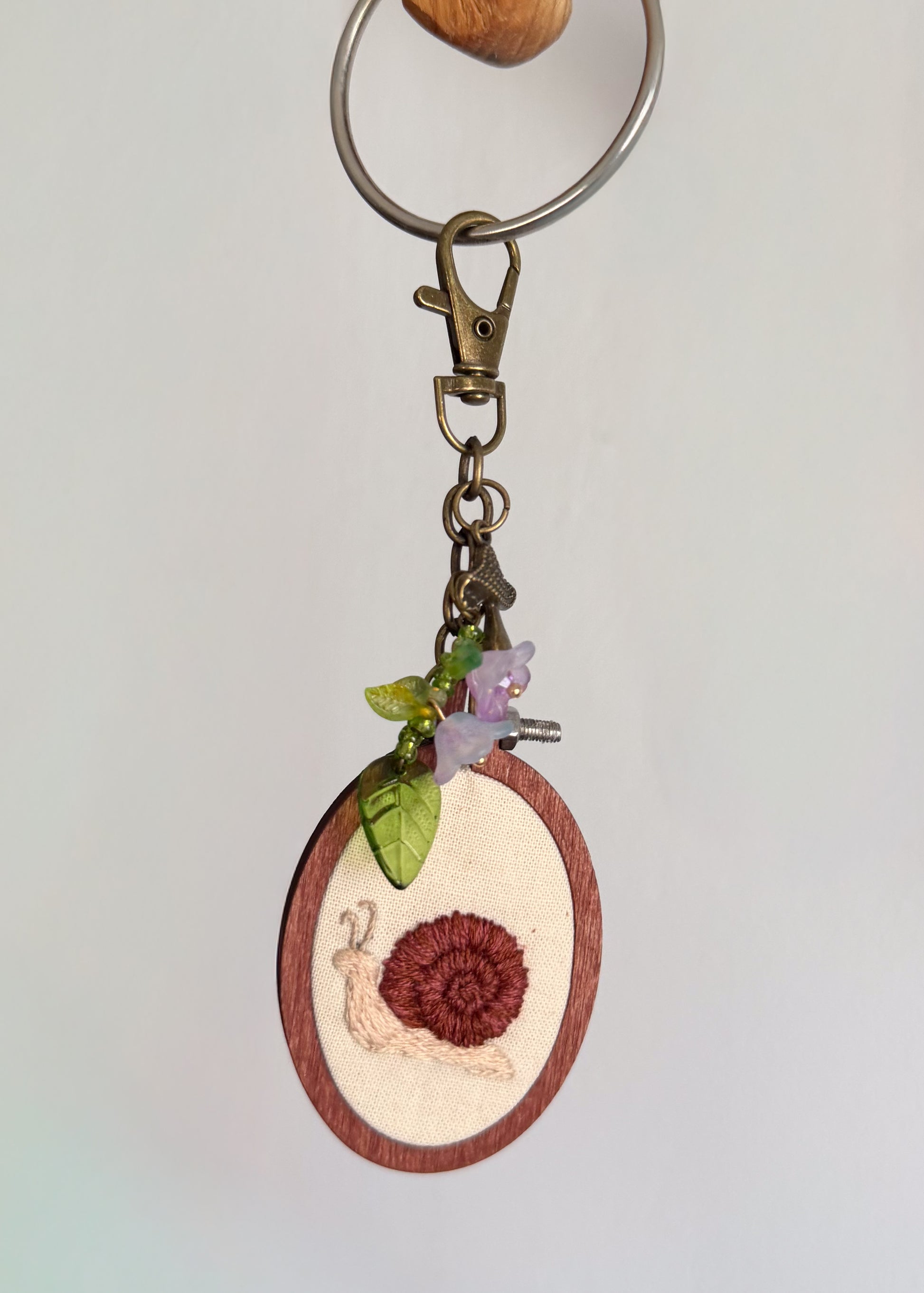 A close-up of a keychain featuring a small oval wooden charm with a snail design, hanging from a metal split ring and clasp against a plain light background.