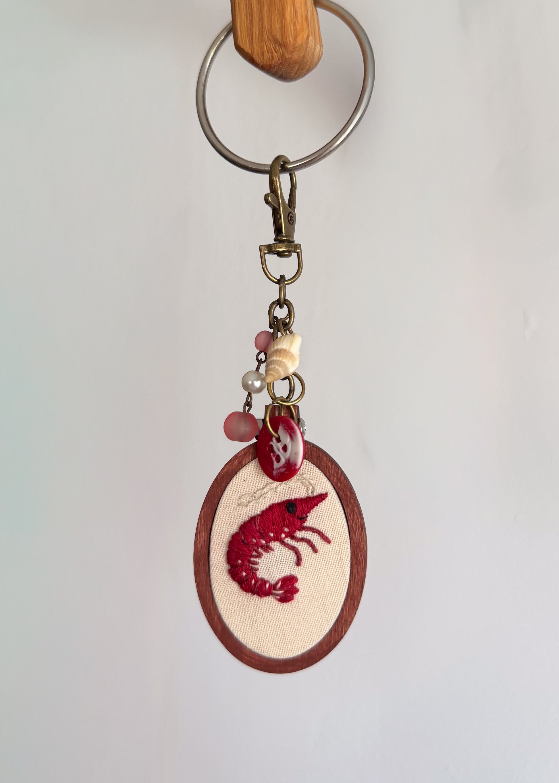 A handmade keychain featuring a circular wooden medallion with red embroidered shrimp, hanging from a metal split ring and clasp.