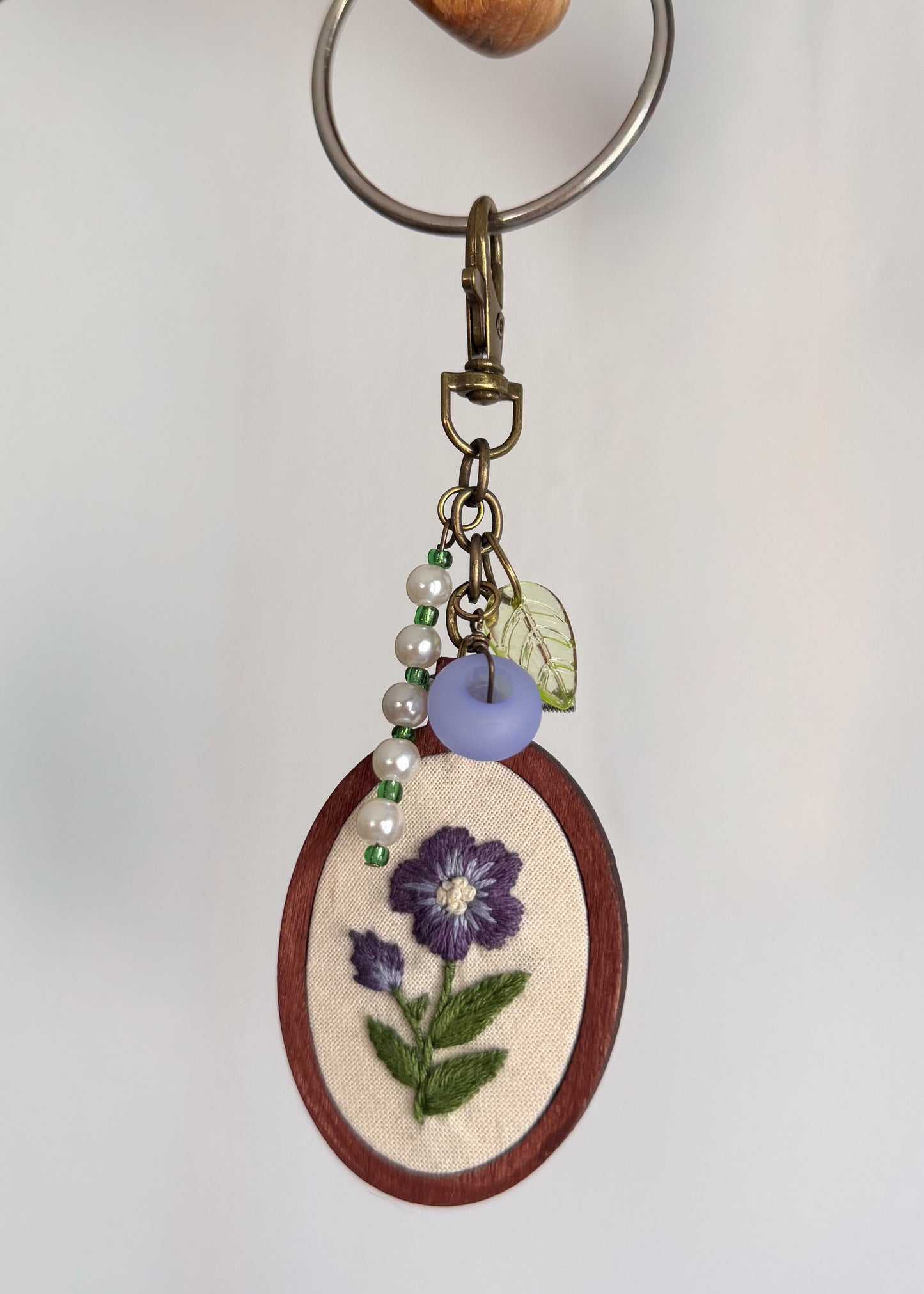 An oval embroidered keychain featuring purple flowers and green leaves on cream fabric, framed in brown wood, with pearl beads, a lavender bead, a small leaf charm, and a metal key ring.
