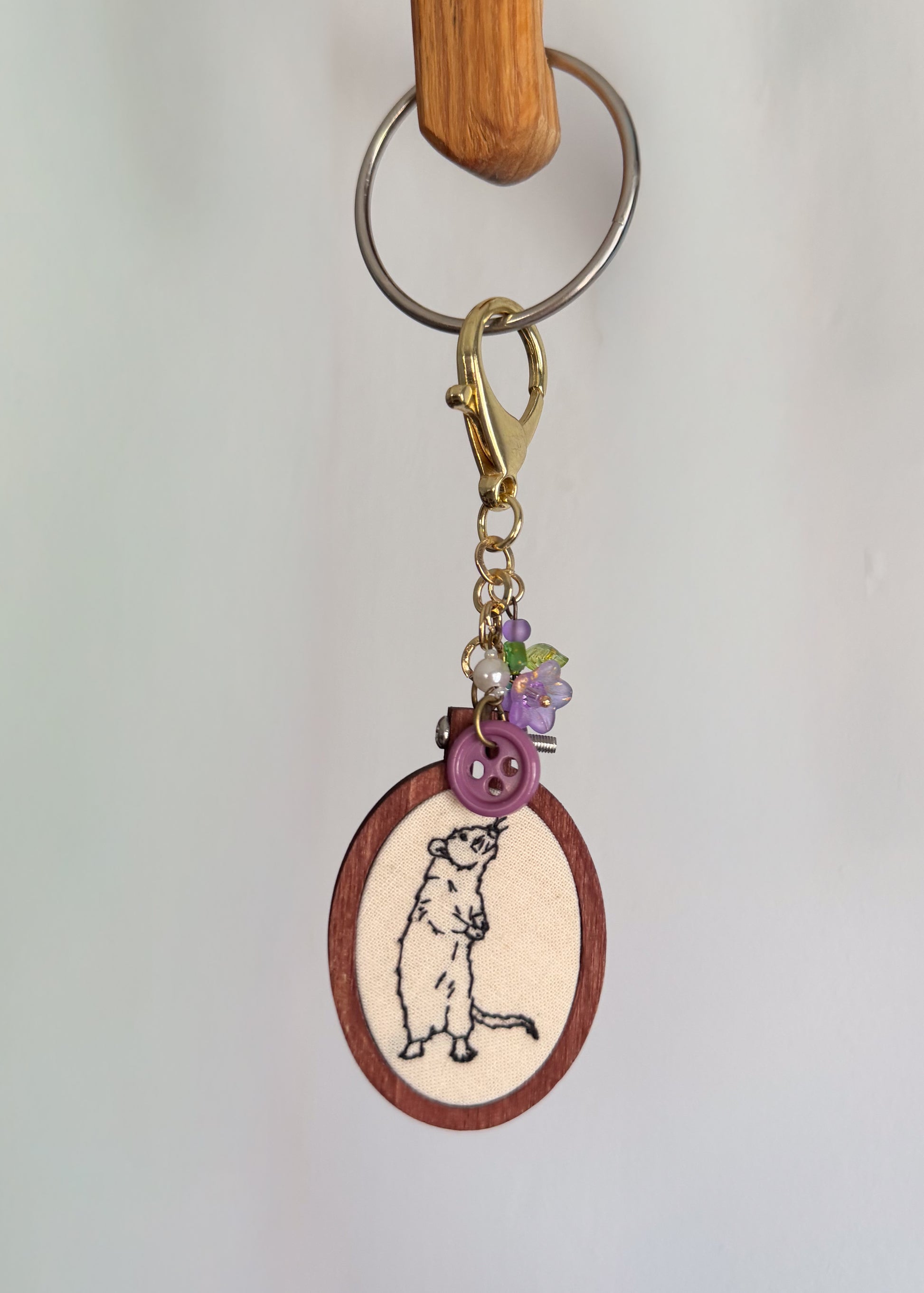 A handmade keychain featuring a wooden oval charm with an embroidered mouse design, adorned with a purple button, purple flower charms, and a gold clasp, hanging from a metal ring on a wooden peg against a white wall.