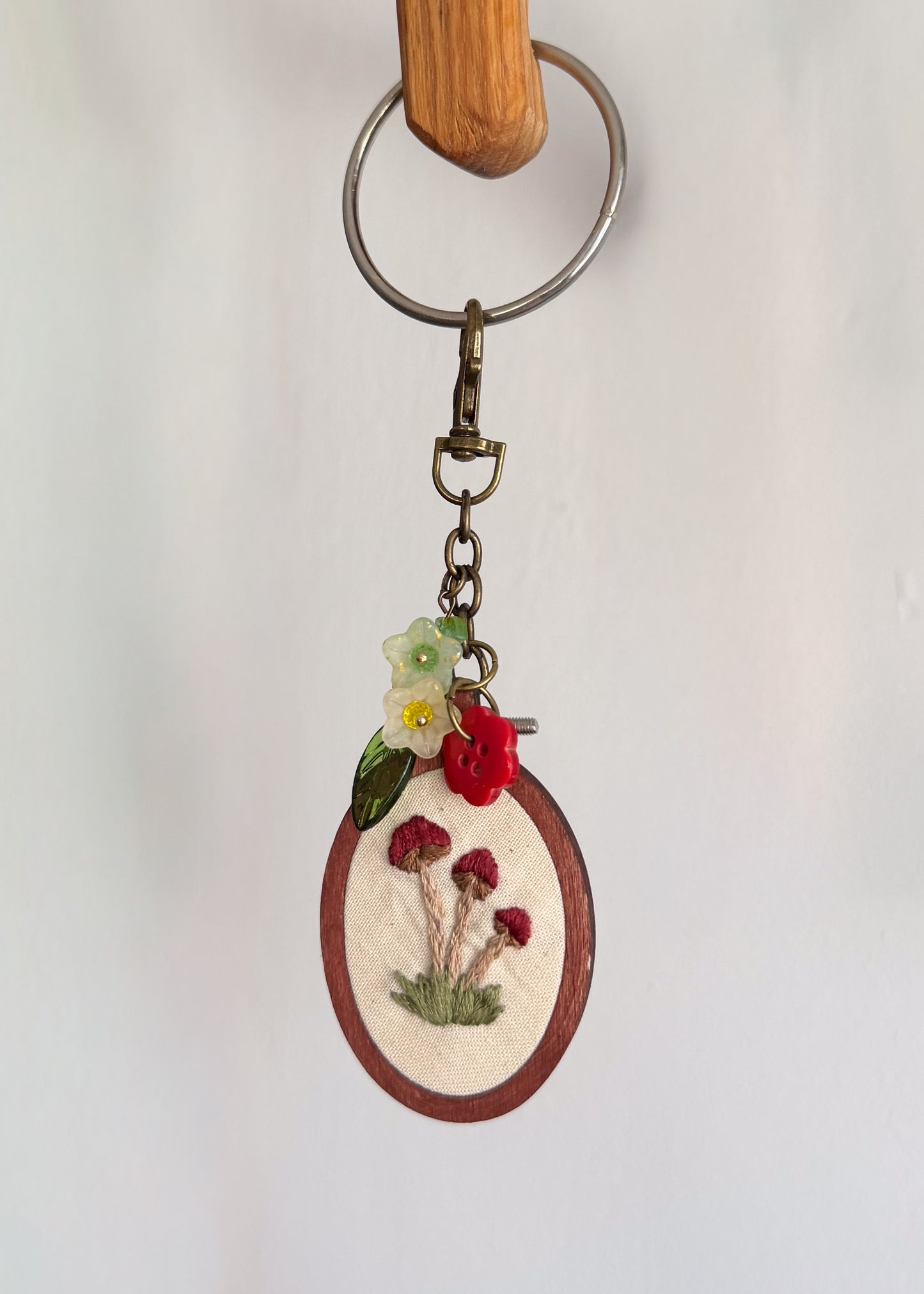 A handmade oval keychain featuring red mushrooms and small white and yellow flowers on a cream fabric background, framed by a brown wooden border, with a silver metal key ring and clasp.