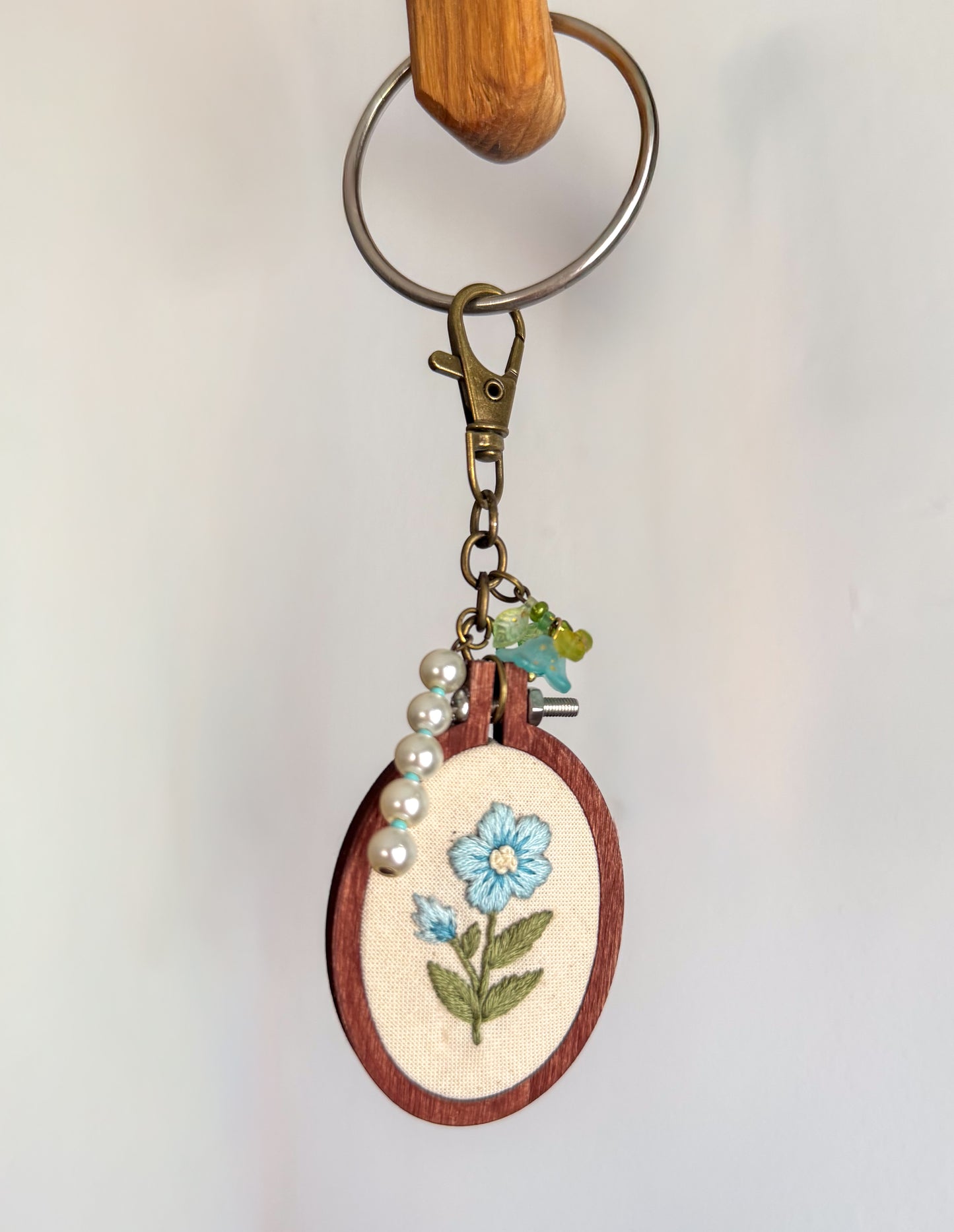 A handmade wooden embroidery hoop keychain featuring a light blue flower with green leaves stitched on cream fabric. The hoop is attached to a brass key ring, clasp, and charm, with a string of white faux pearls draped on the side.
