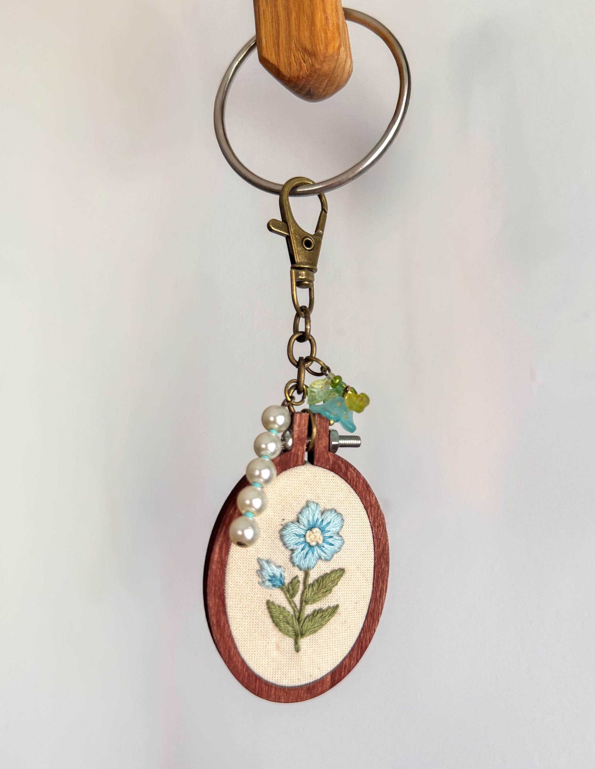 A handmade wooden embroidery hoop keychain featuring a light blue flower with green leaves stitched on cream fabric. The hoop is attached to a brass key ring, clasp, and charm, with a string of white faux pearls draped on the side.
