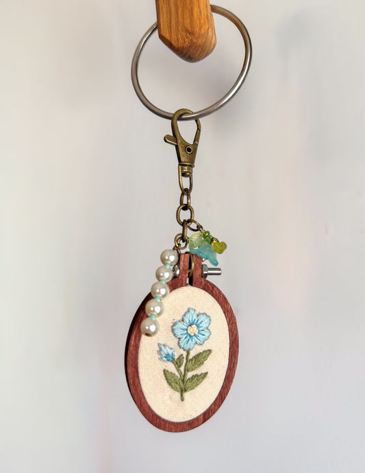 A handmade wooden embroidery hoop keychain featuring a light blue flower with green leaves stitched on cream fabric. The hoop is attached to a brass key ring, clasp, and charm, with a string of white faux pearls draped on the side.