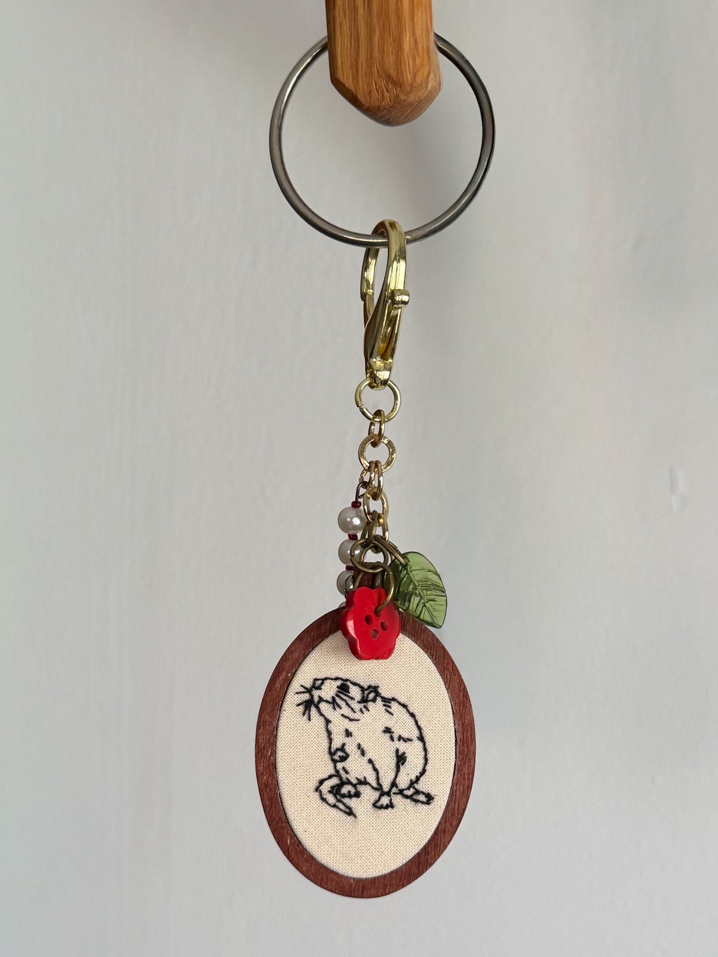 A handmade keychain featuring an oval wooden charm with a cream-colored fabric insert embroidered with a outline of a rat. The charm is decorated with a small red button and a green leaf, attached to a gold-tone clasp and a metal split ring.