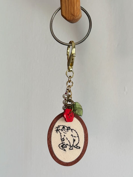 A handmade keychain featuring an oval wooden charm with a cream-colored fabric insert embroidered with a outline of a rat. The charm is decorated with a small red button and a green leaf, attached to a gold-tone clasp and a metal split ring.