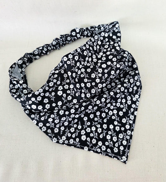 A black fabric hair bandana with a white floral pattern, laid flat on a light beige fabric background.