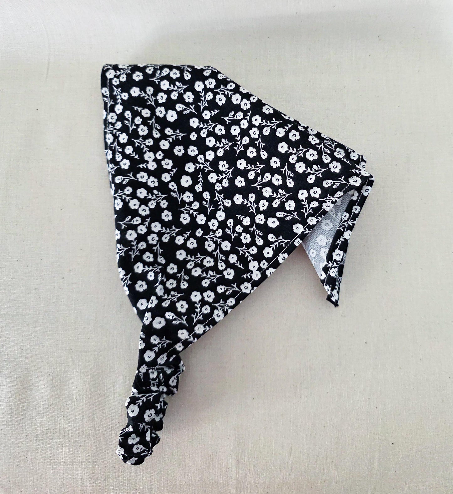 A black hair bandana with a white floral pattern and elastic band laid flat on a light beige fabric background.