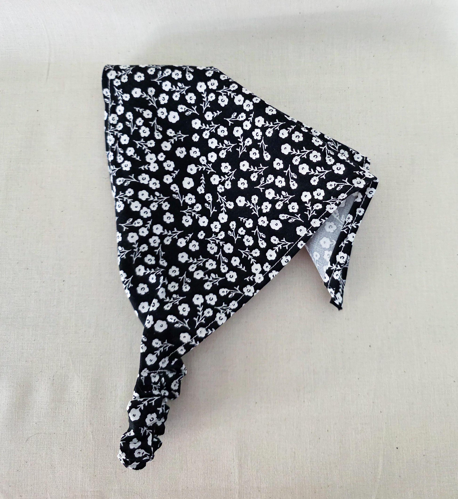 A black hair bandana with a white floral pattern and elastic band laid flat on a light beige fabric background.