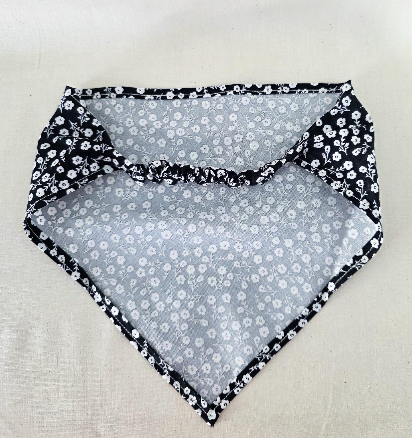 A triangular hair bandana laid flat on a light beige fabric background. The fabric features a black and white floral pattern 