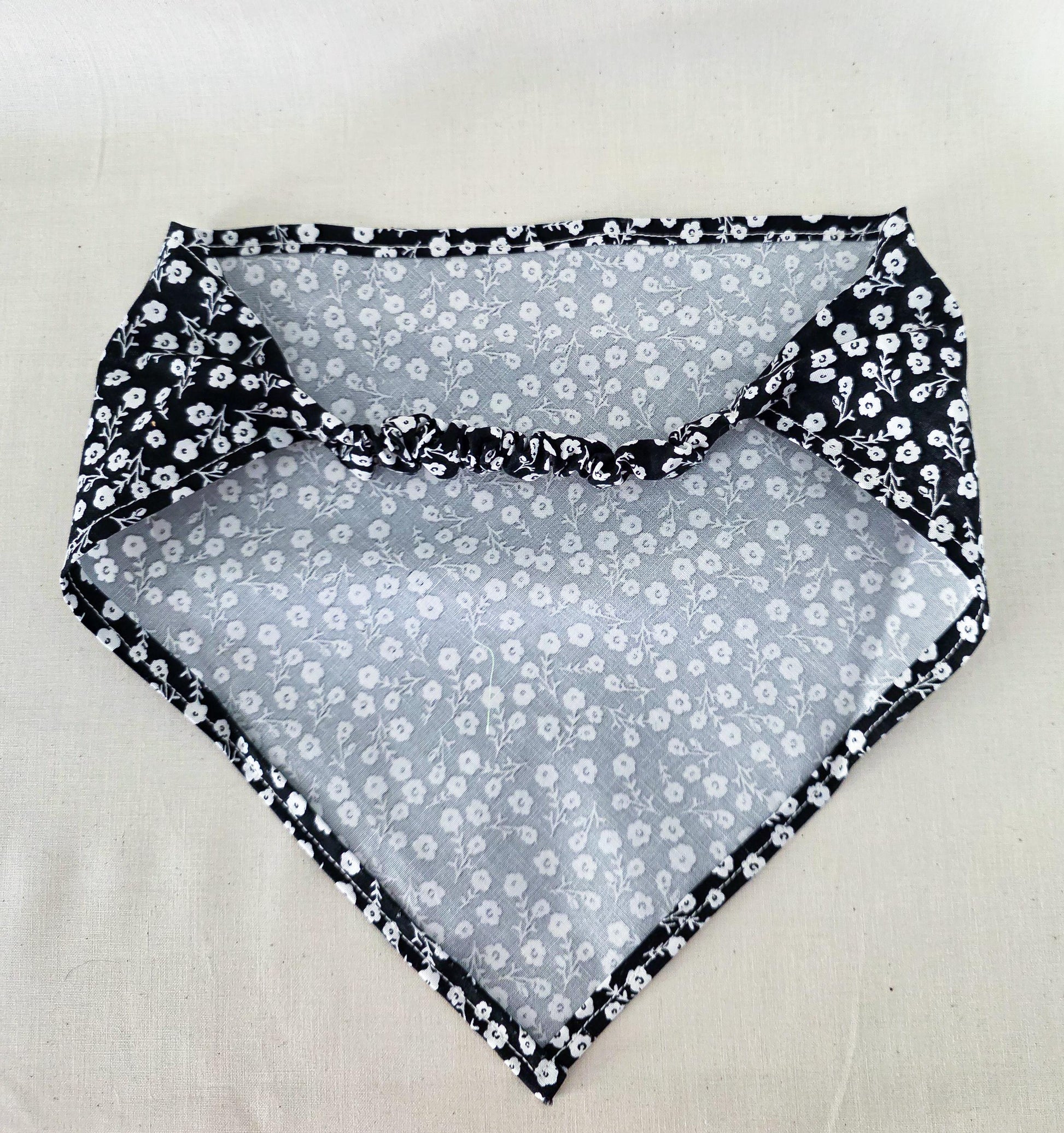 A triangular hair bandana laid flat on a light beige fabric background. The fabric features a black and white floral pattern 