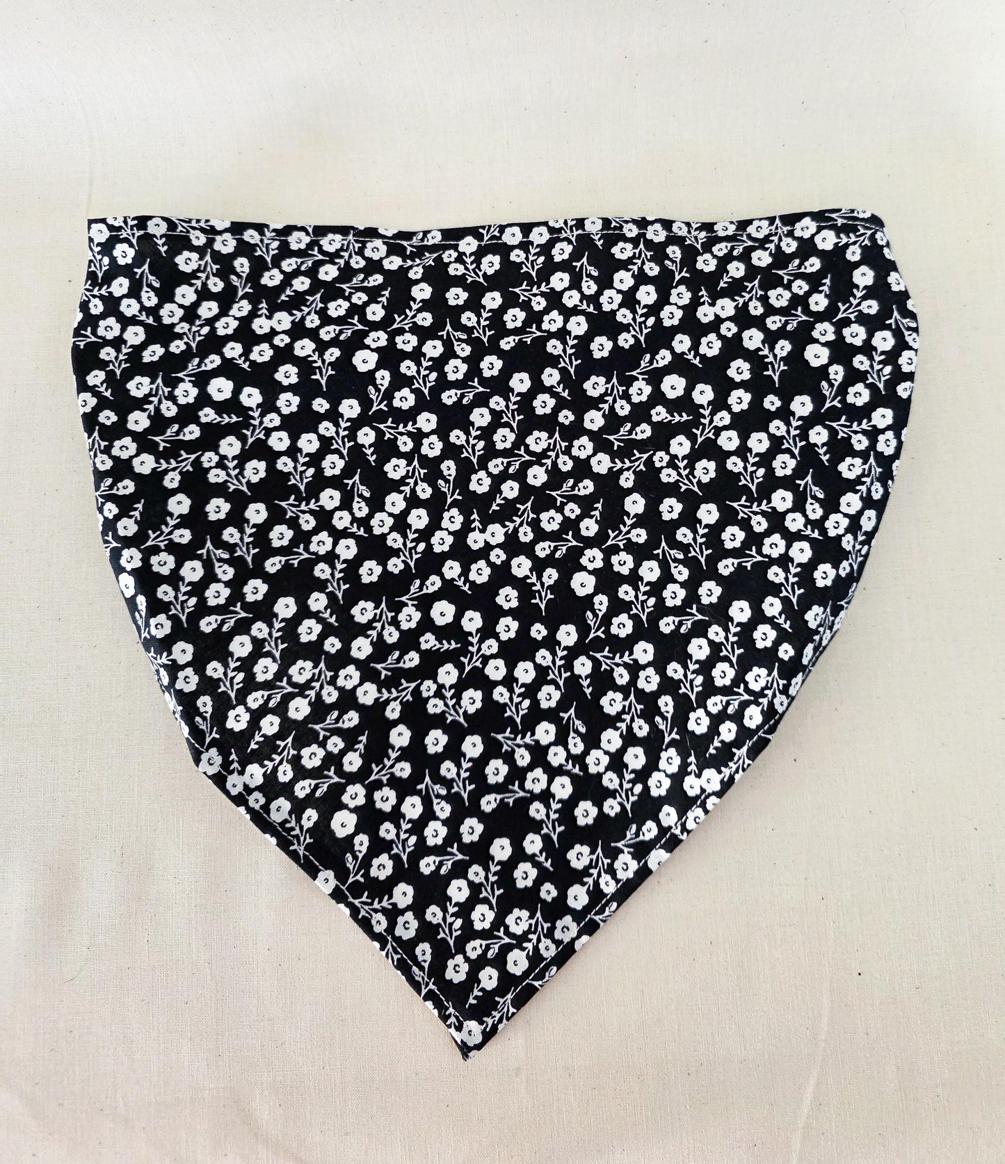 A black triangular hair bandana fabric piece with a dense white floral print, laid flat on a light beige fabric background.