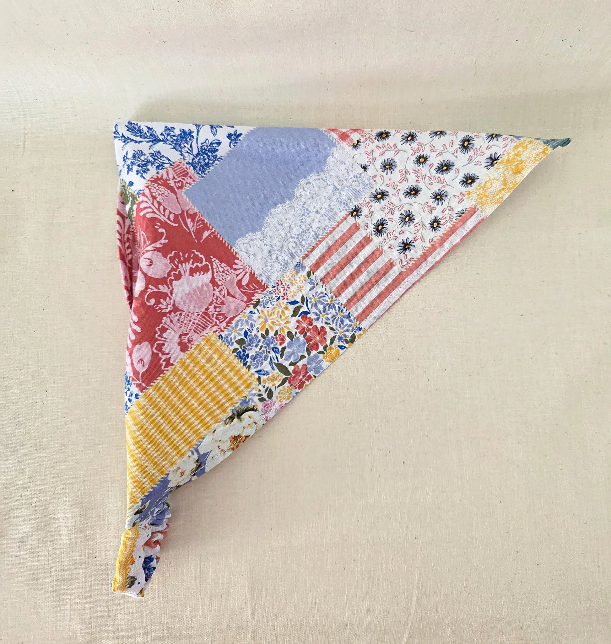 A triangular faux patchwork hair bandana made of multiple fabric patterns including floral, stripes, lace, and solid colors, displayed on a neutral beige fabric background.