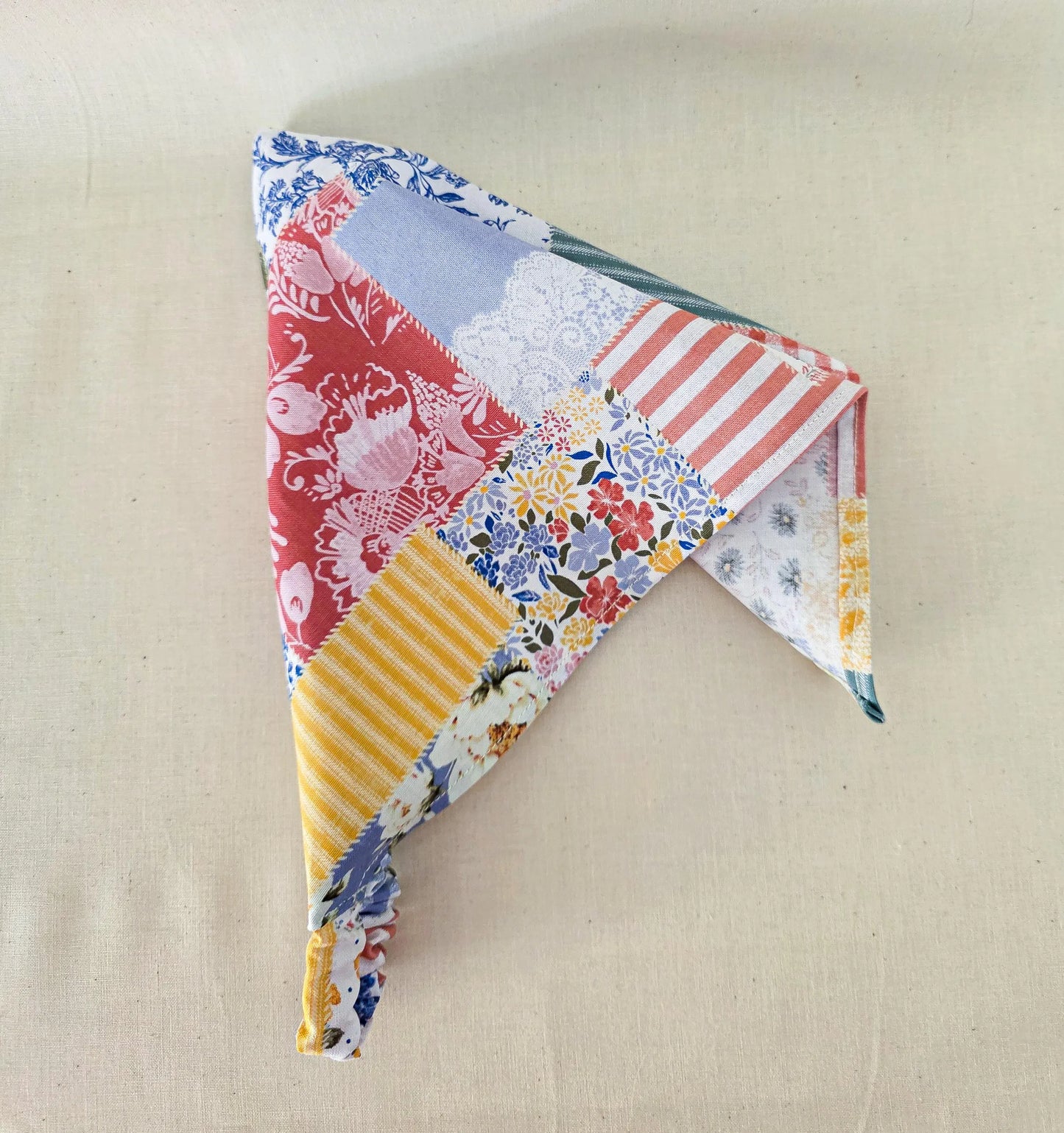 A triangular faux patchwork bandana made from multiple fabric prints, including floral, striped, and solid patterns in red, blue, yellow, white, and green, displayed on a neutral beige linen background.