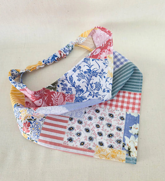 A handmade faux patchwork hair bandana with a gathered elastic strap, featuring various colorful fabric prints including blue florals, red gingham, yellow stripes, and various florals, displayed on a neutral beige fabric background.