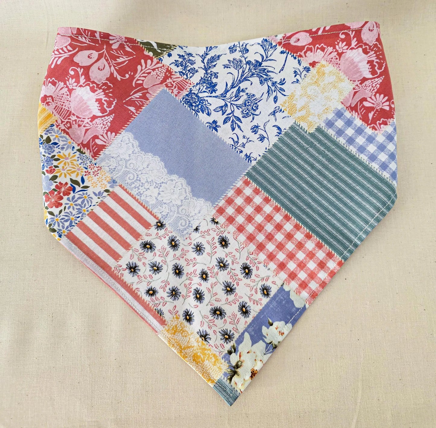 A handmade triangular hair bandana made from a patchwork of various fabric prints including gingham, floral, stripes, and lace, displayed flat on a beige linen background.
