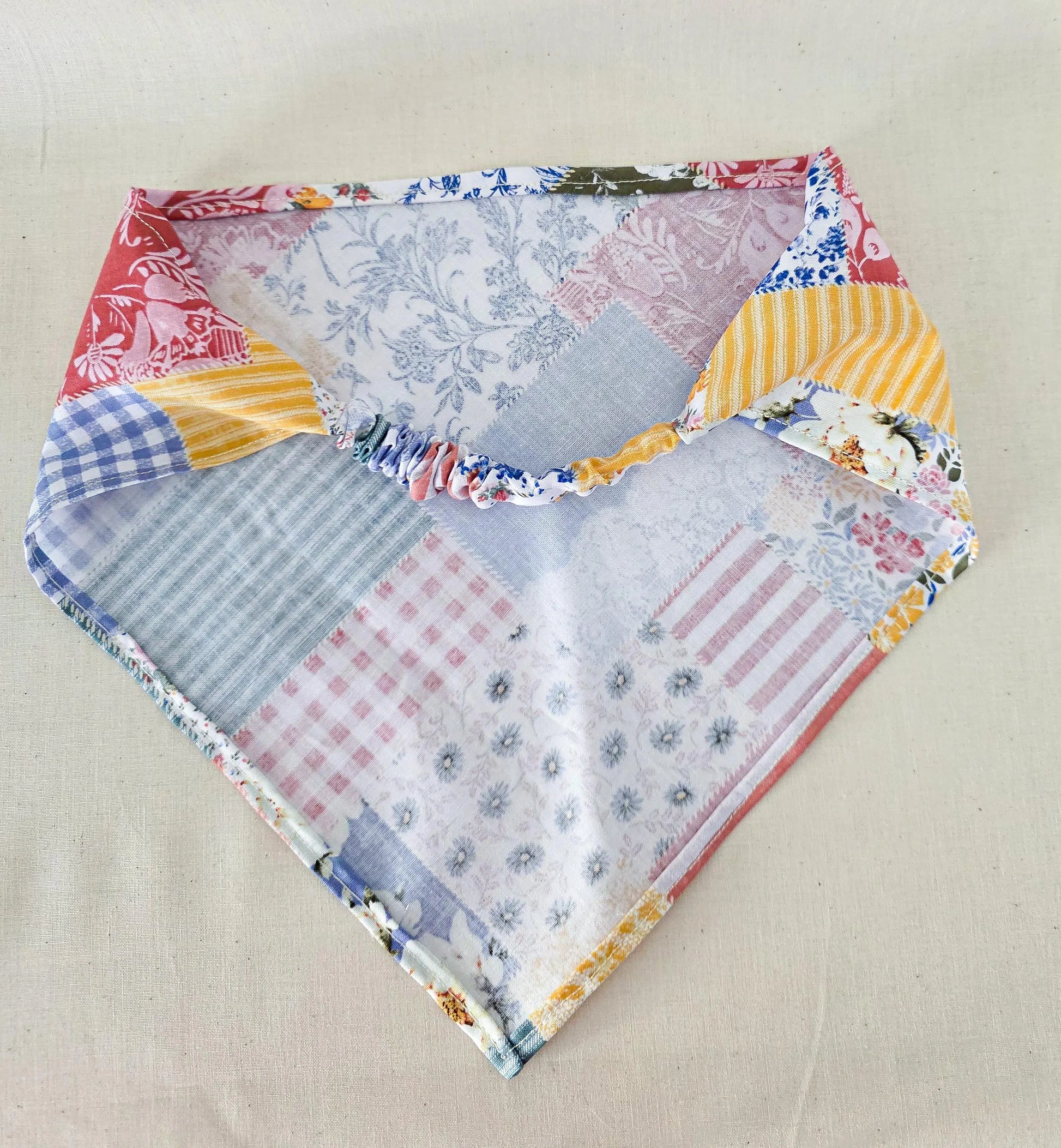 A handmade triangular hair bandana  with a colorful faux patchwork print of various fabrics, laid flat on a neutral beige fabric background.