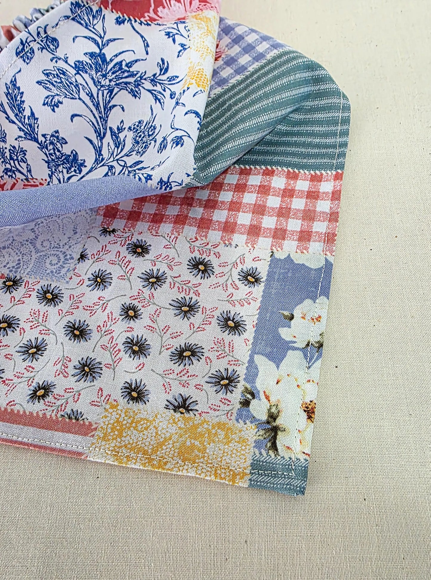 A faux patchwork fabric piece with multiple printed and striped sections, including blue floral, red gingham, black-and-white floral, and yellow patterned areas, laid on a light beige textured fabric background.