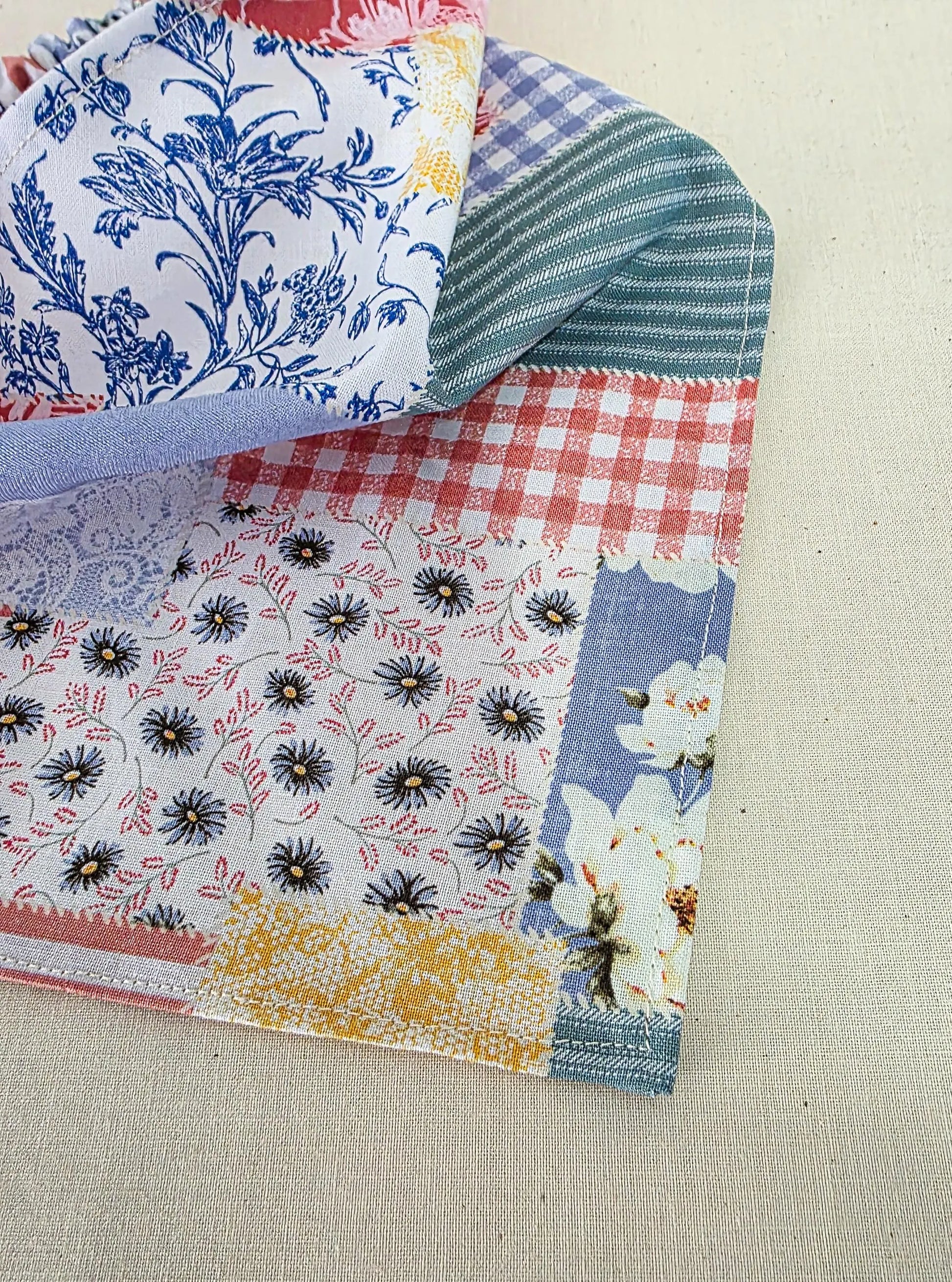 A faux patchwork fabric piece with multiple printed and striped sections, including blue floral, red gingham, black-and-white floral, and yellow patterned areas, laid on a light beige textured fabric background.