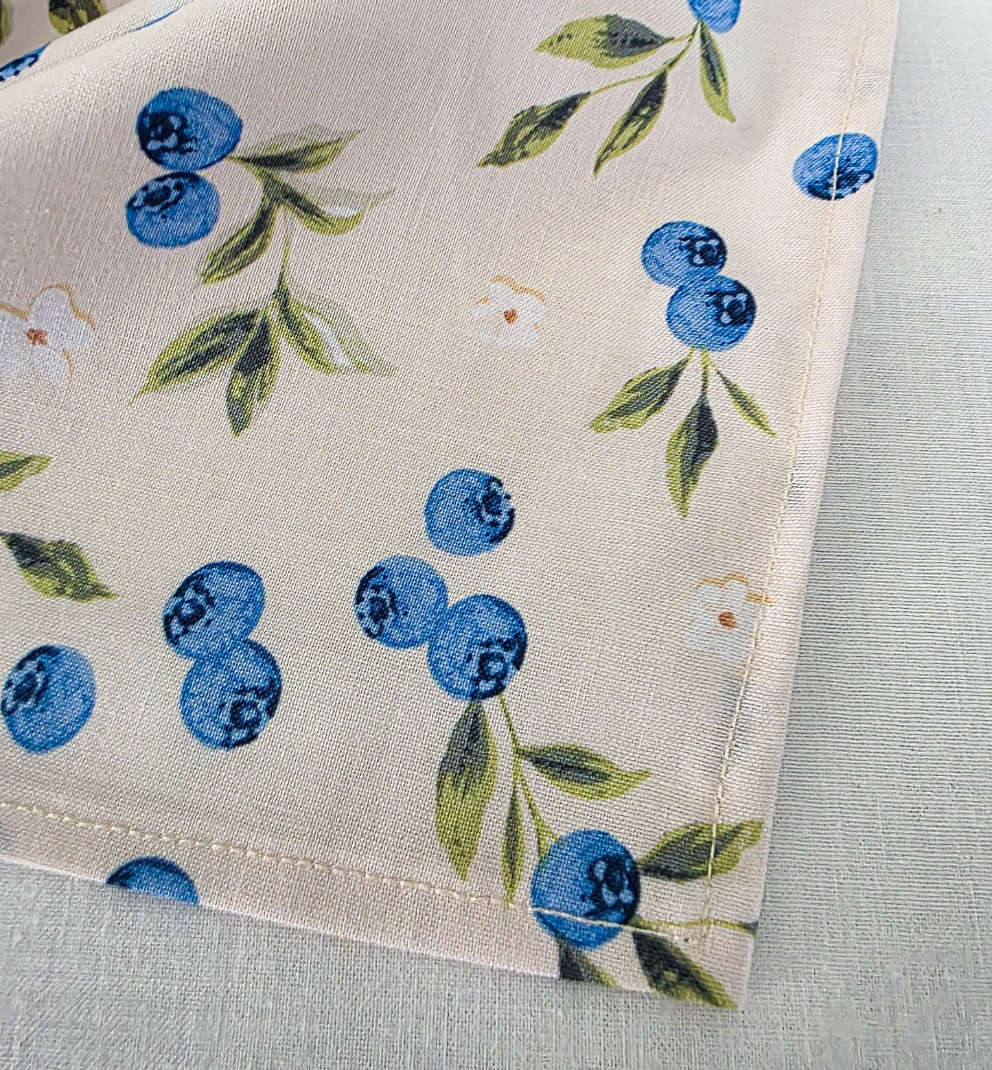 A close-up of a cream-colored fabric bandana featuring a printed pattern of blue blueberries, green leaves, and small white flowers, laid flat on a textured light surface.