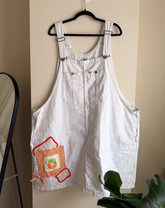 White overall dress with a decorative patch on a hanger against a beige wall.