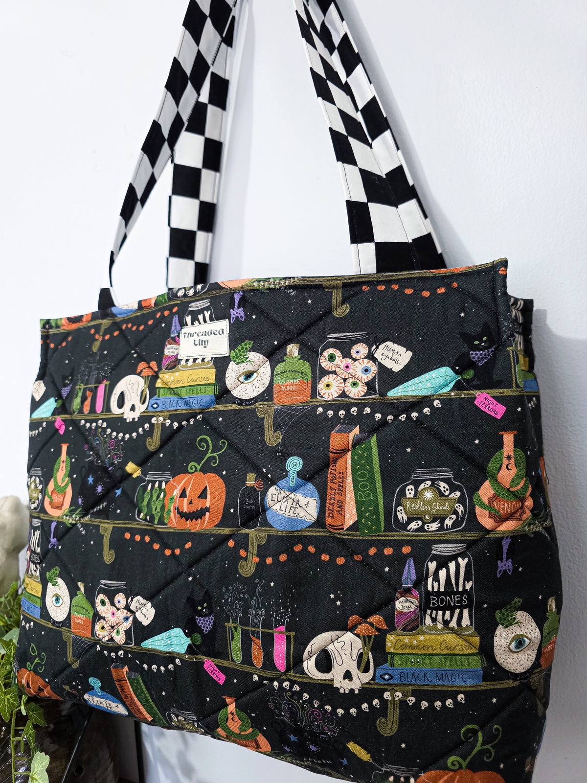 Large quilted tote bag with a witchy halloween bookshelf patterned fabric and black and white checkered straps 