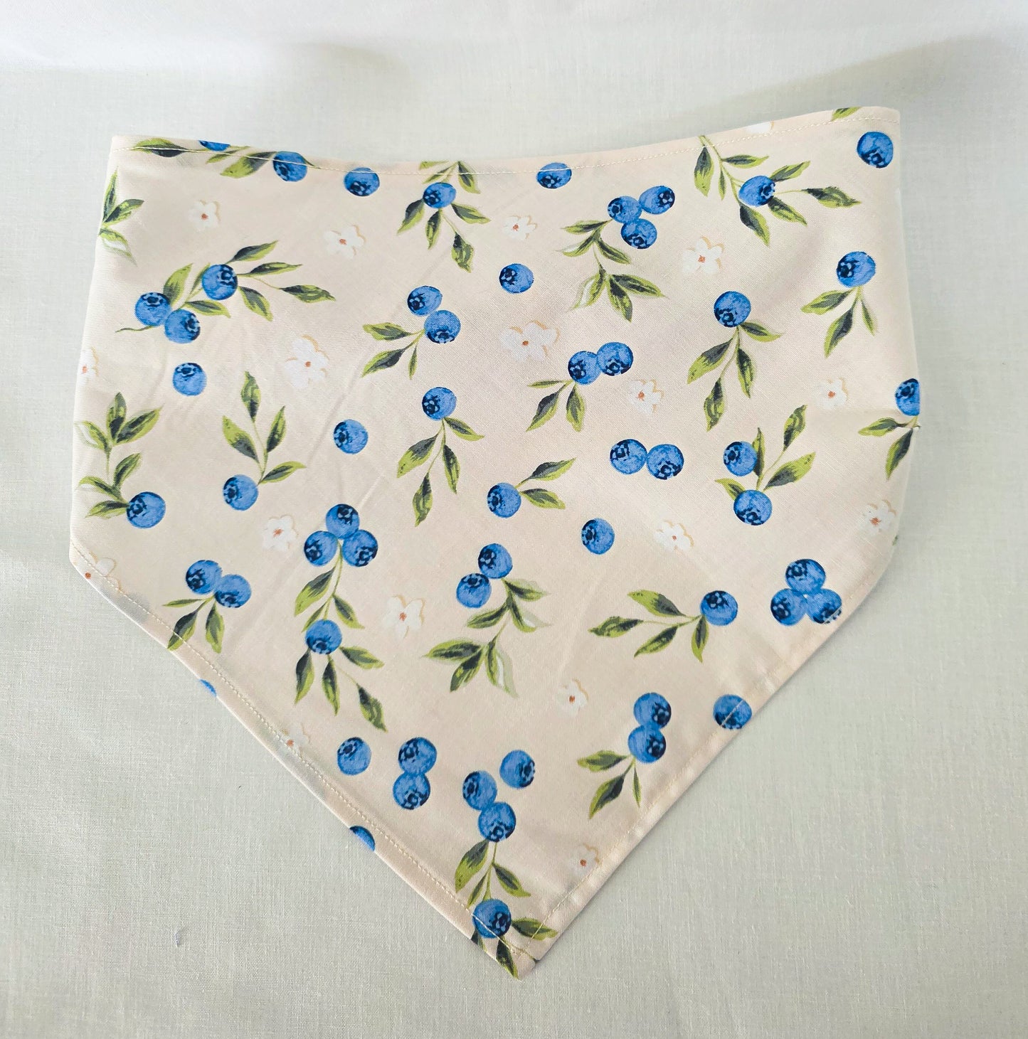 A triangular hair bandana with a blueberry and green leaf pattern on a light cream background, laid flat on a light fabric surface.