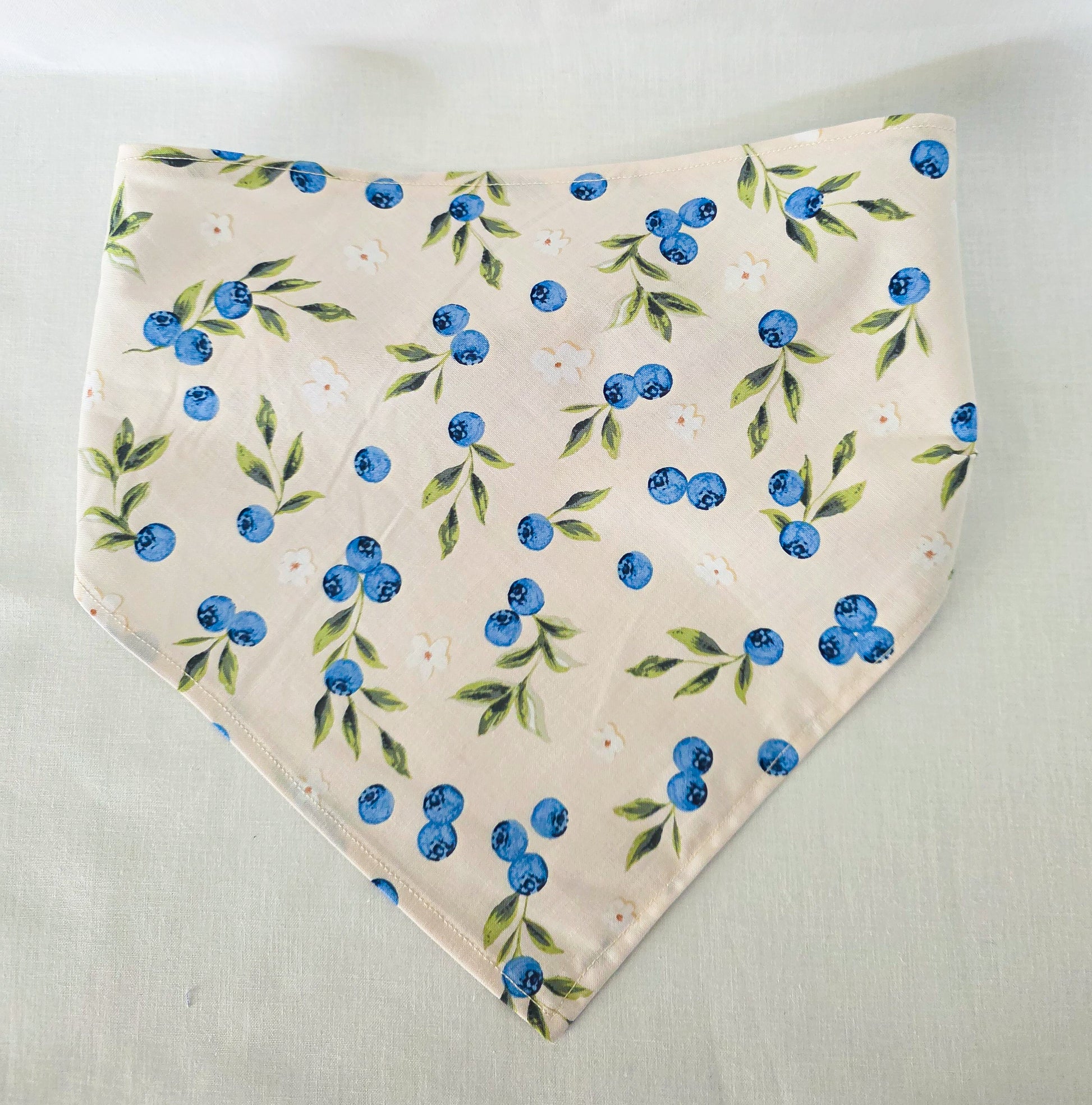 A triangular hair bandana with a blueberry and green leaf pattern on a light cream background, laid flat on a light fabric surface.