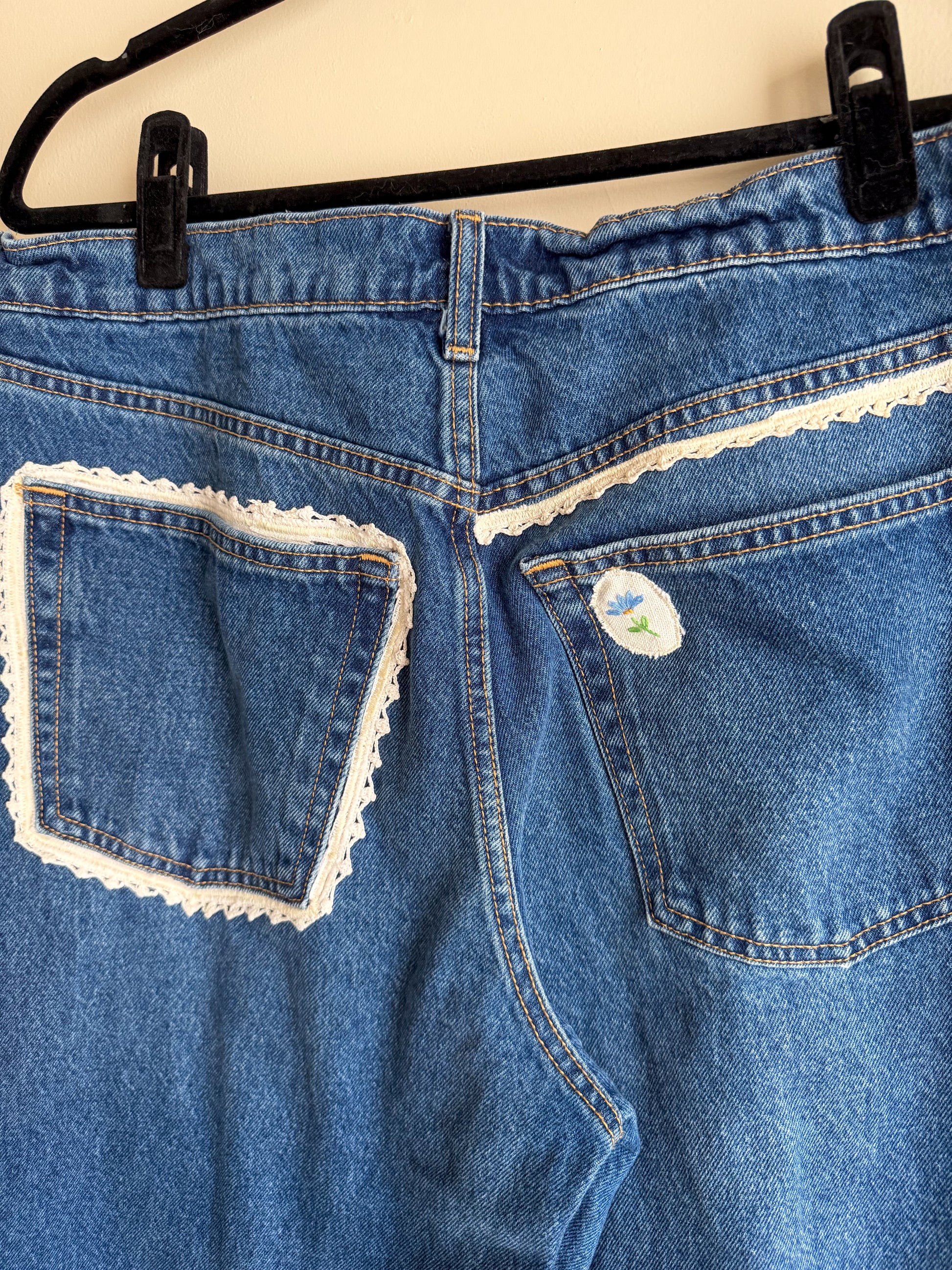 Blue denim jeans with lace trim on pockets, hanging on a black hanger.